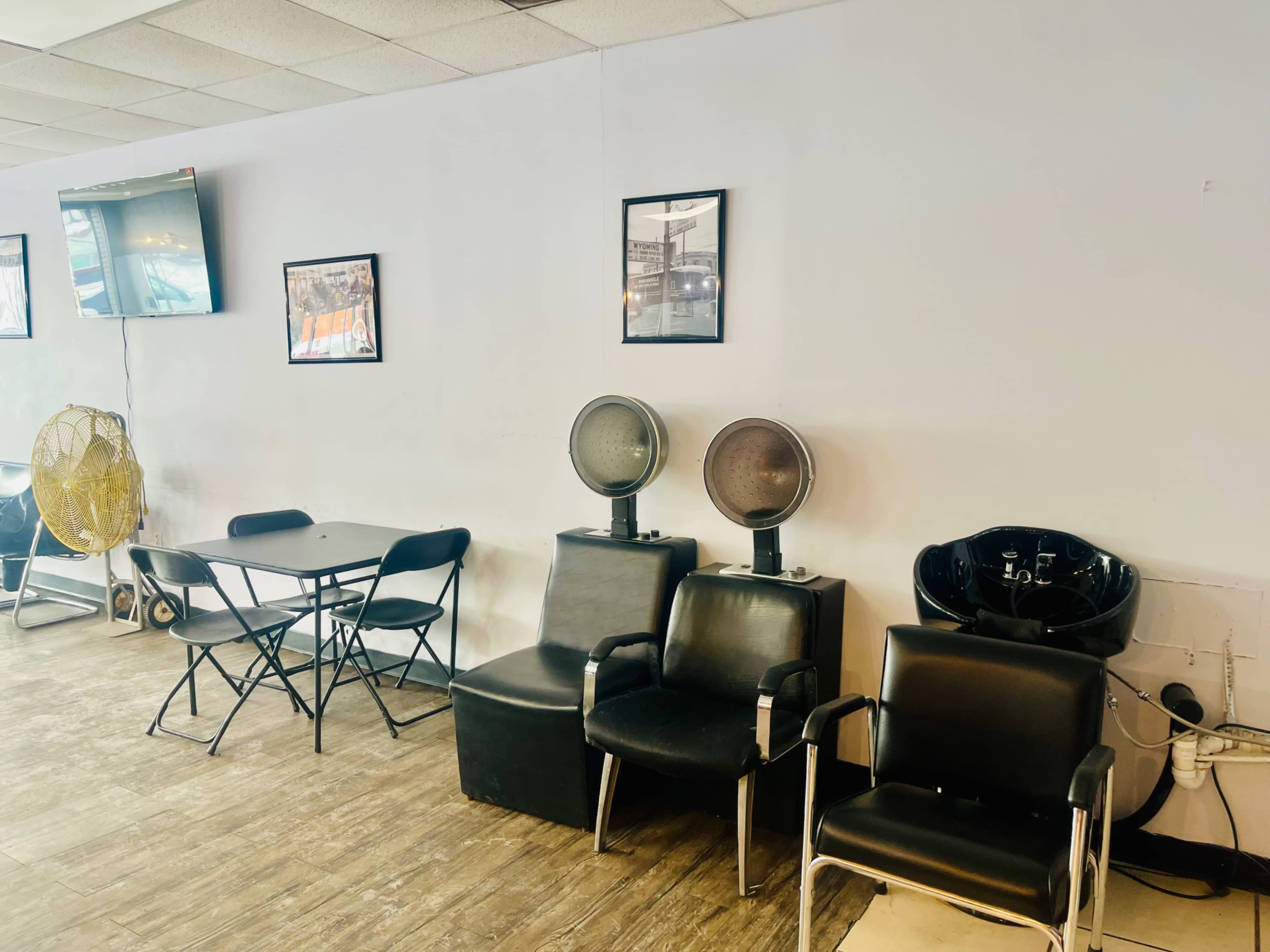 The image shows a hair salon interior featuring black chairs, a table, hair dryers, and framed pictures on the wall.