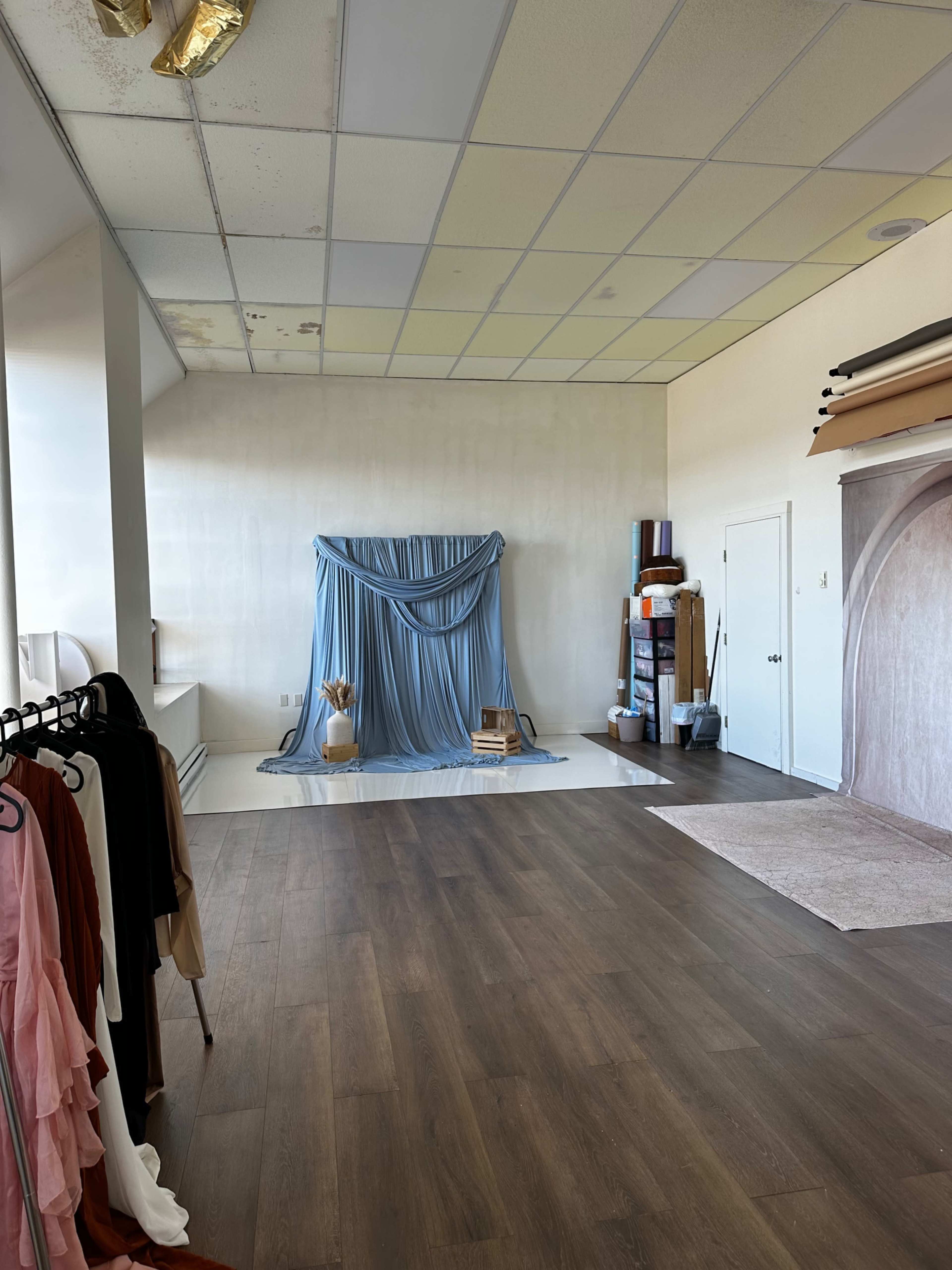 The image shows an empty room with wooden flooring, a draped blue backdrop in the corner, and various items including clothing racks and storage units along the walls.