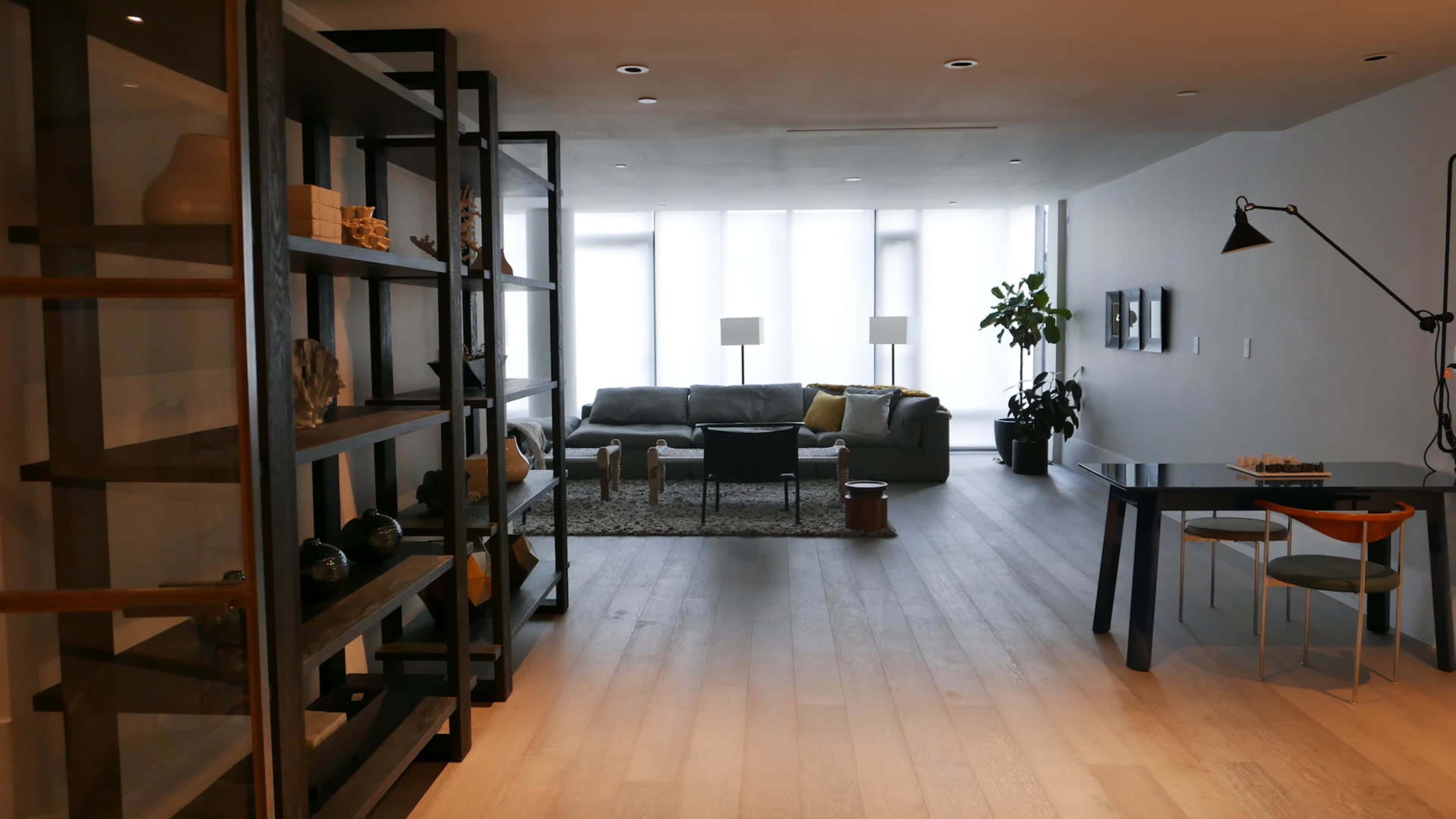 The image shows a modern living space with a grey sofa, a dining table, and large windows that allow natural light to fill the room.