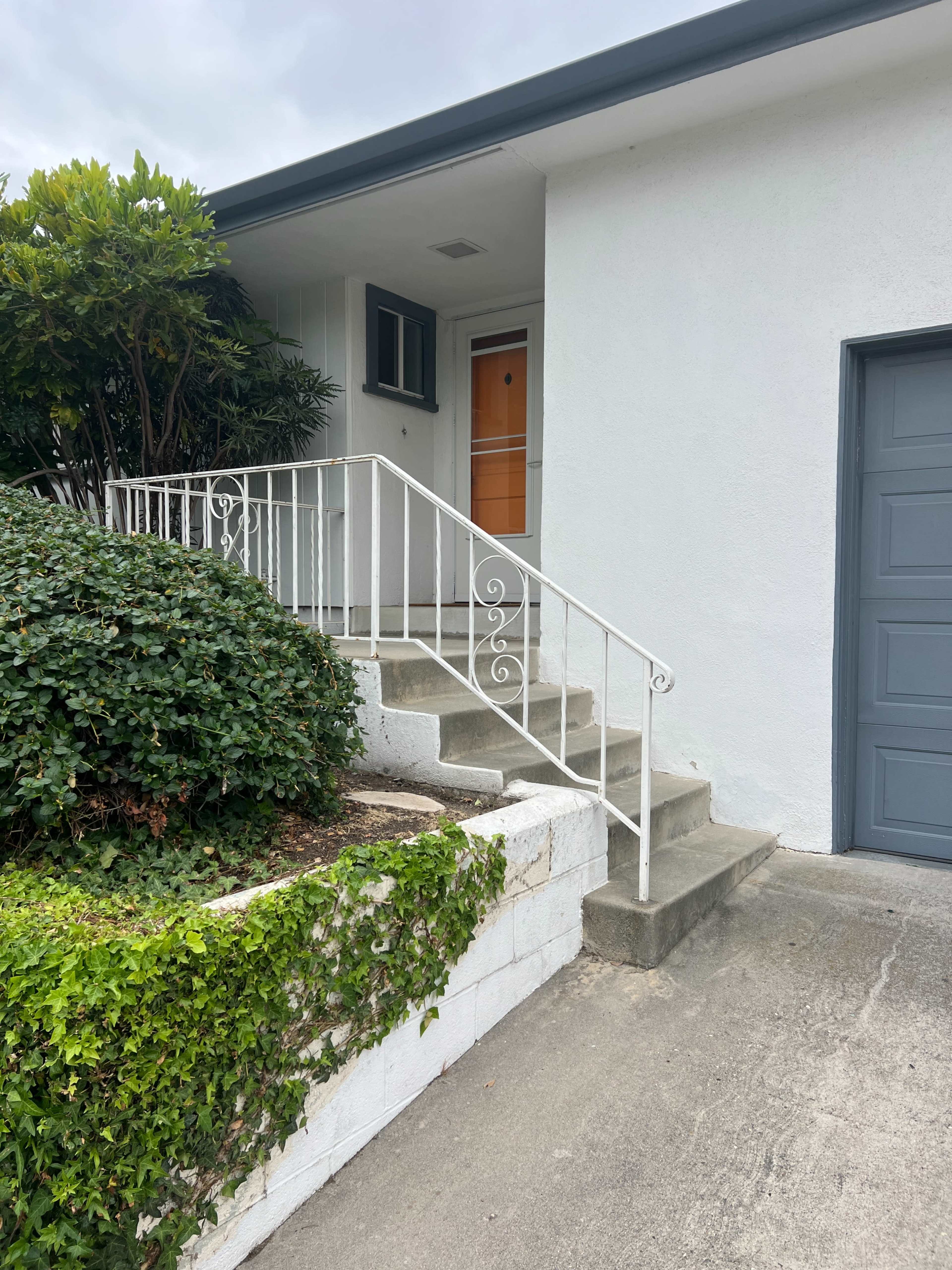 A set of concrete steps leads up to a white house with a metal railing and a door featuring a window, surrounded by green shrubs.