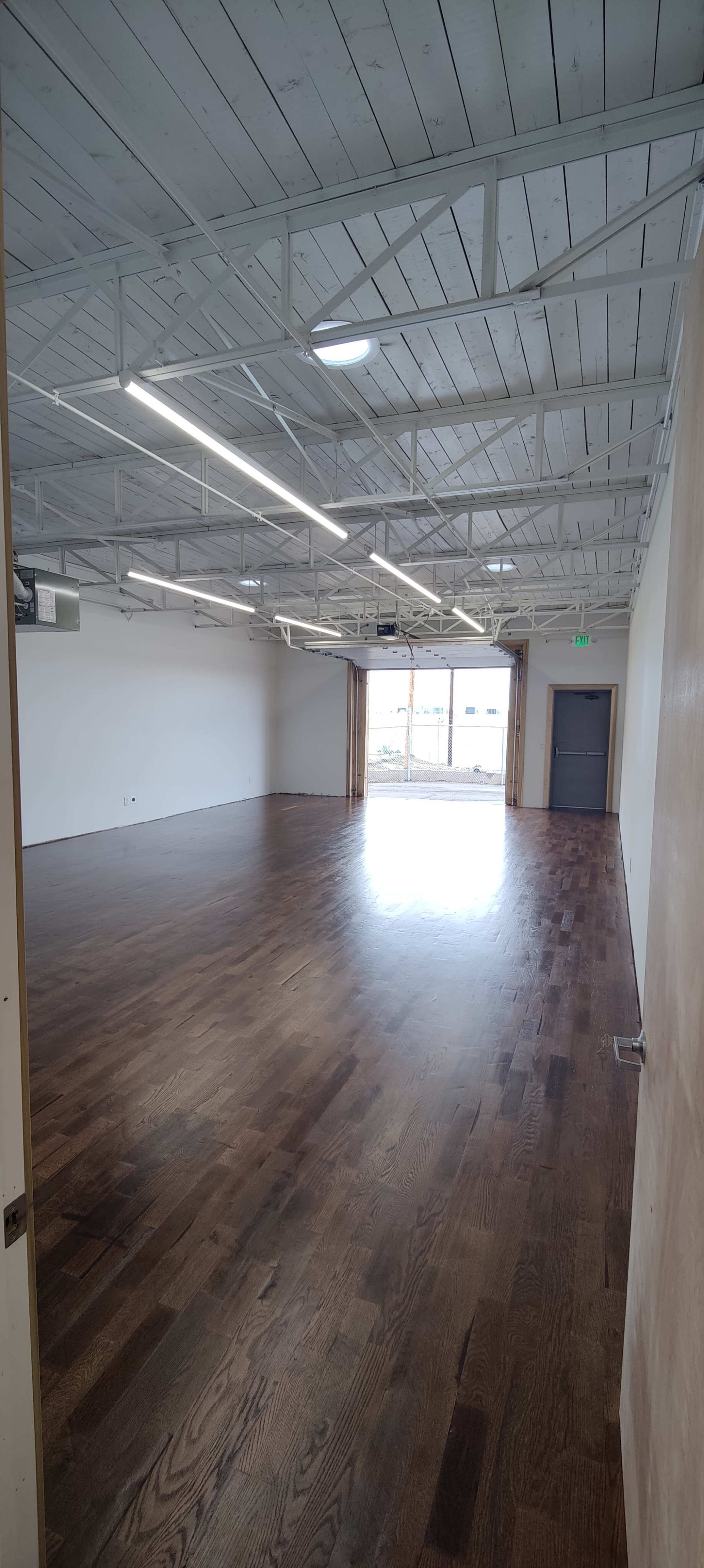 The image shows a spacious indoor room with polished wooden floors and large windows at one end, allowing natural light to enter.