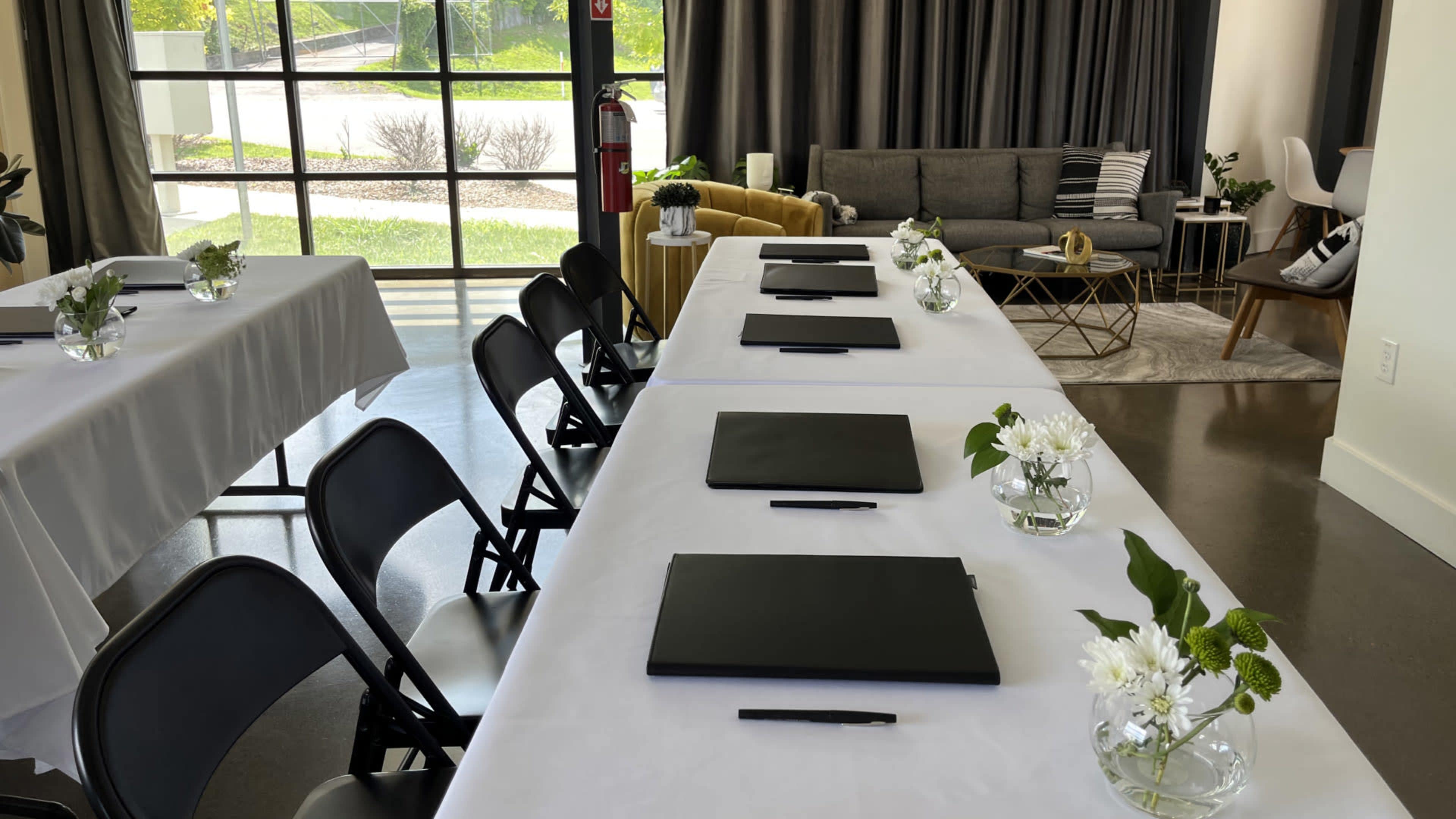 A spacious meeting room features several white-covered tables arranged with black folders and small vases of flowers, set against a backdrop of large windows and modern seating.