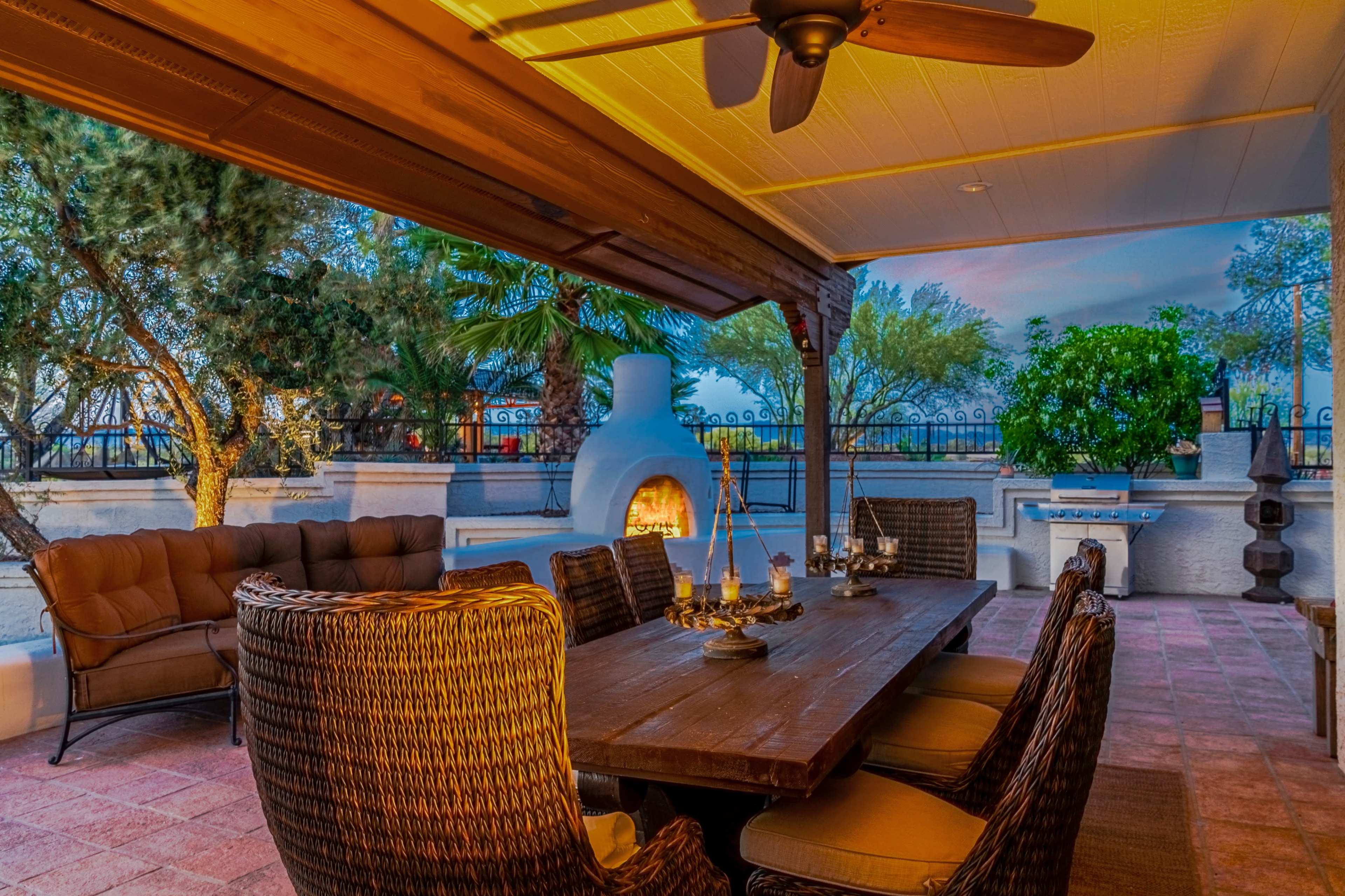 The image depicts a shaded outdoor patio area featuring a wooden dining table surrounded by wicker chairs, a cozy fireplace, and lush greenery in the background.