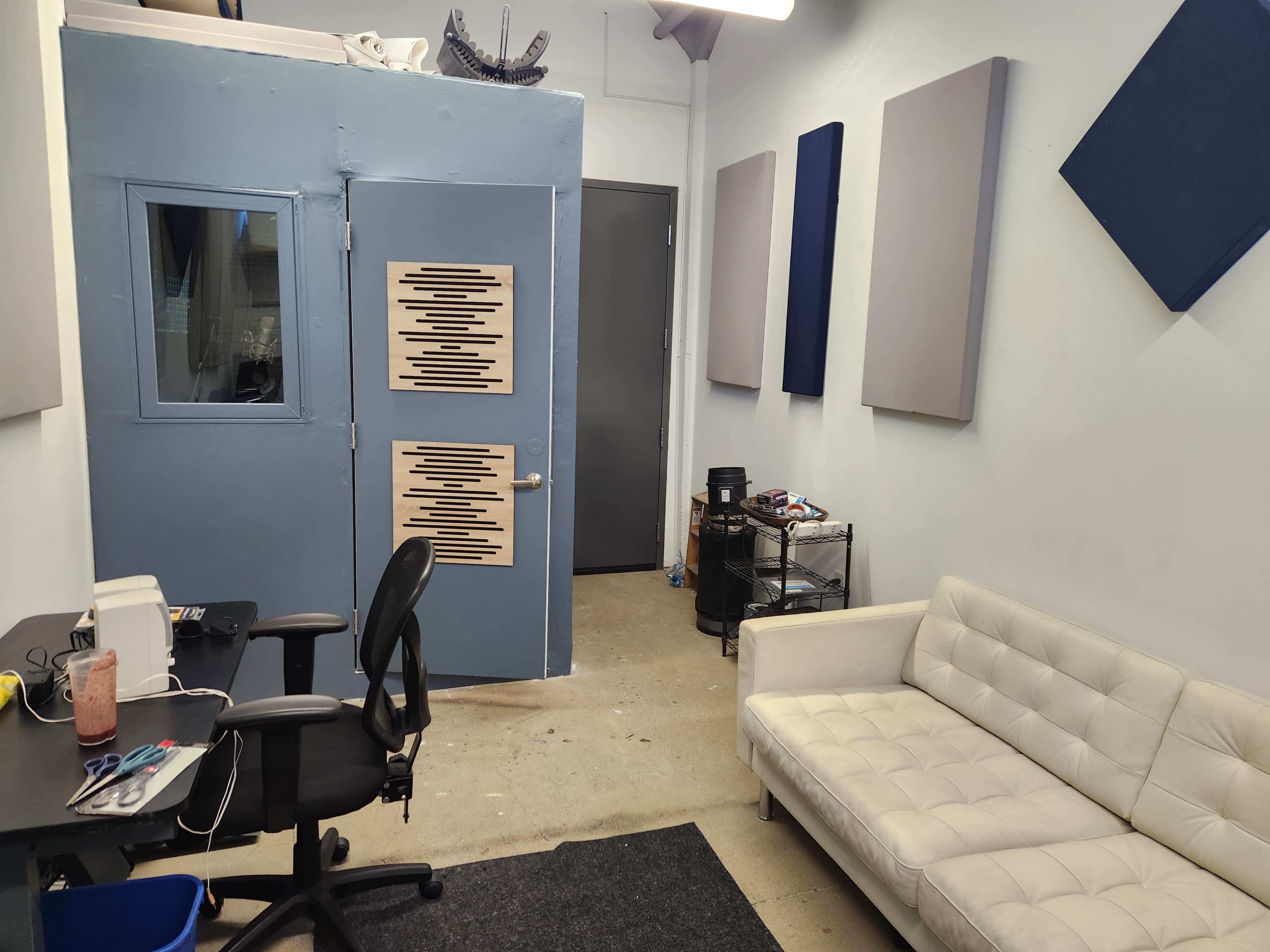A small office or studio space featuring a white couch, a desk with a chair, and a soundproof booth with textured wood panels on the door.