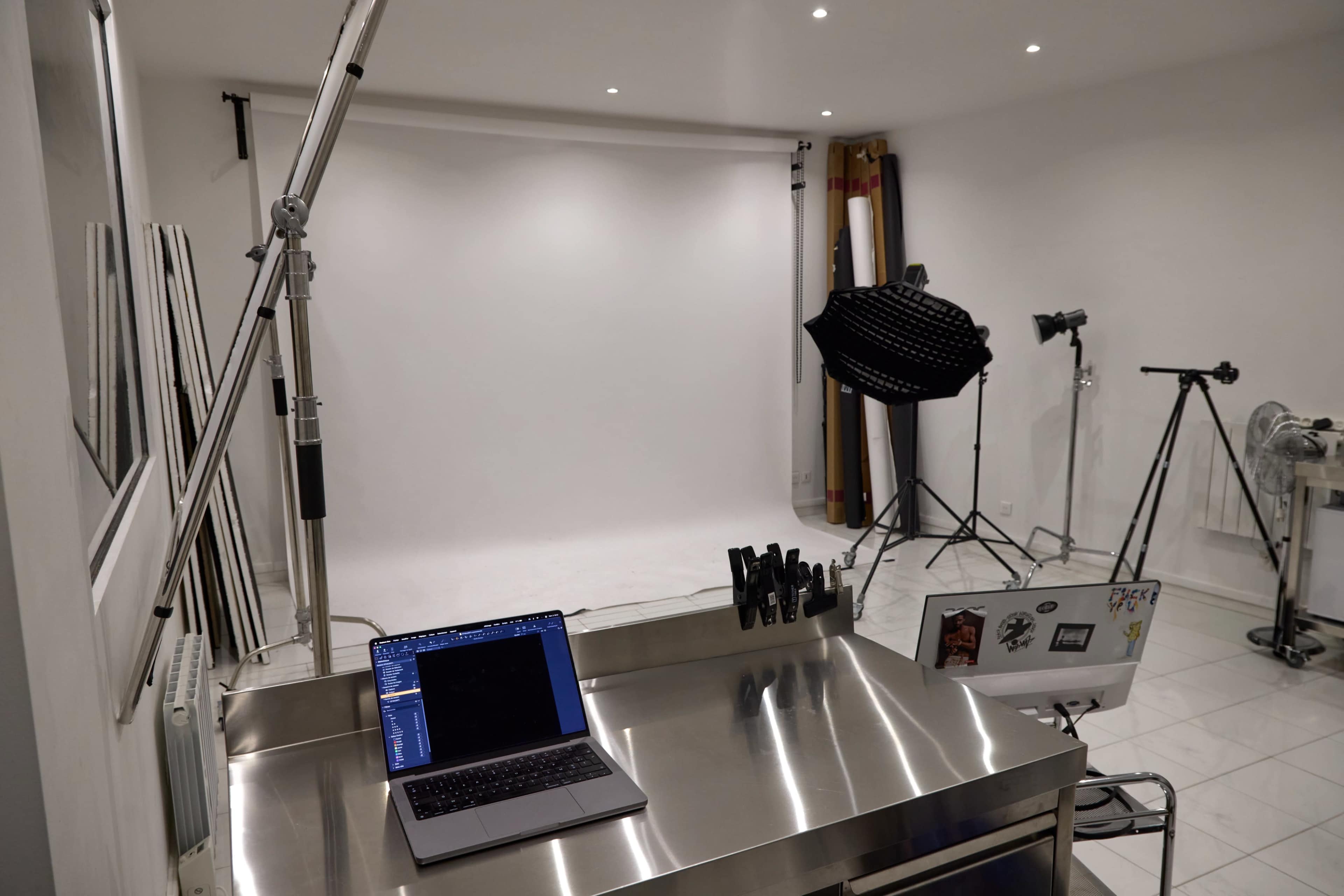 The image shows a photography studio with a blank backdrop, a laptop on a stainless steel table, and various lighting equipment arranged in the background.