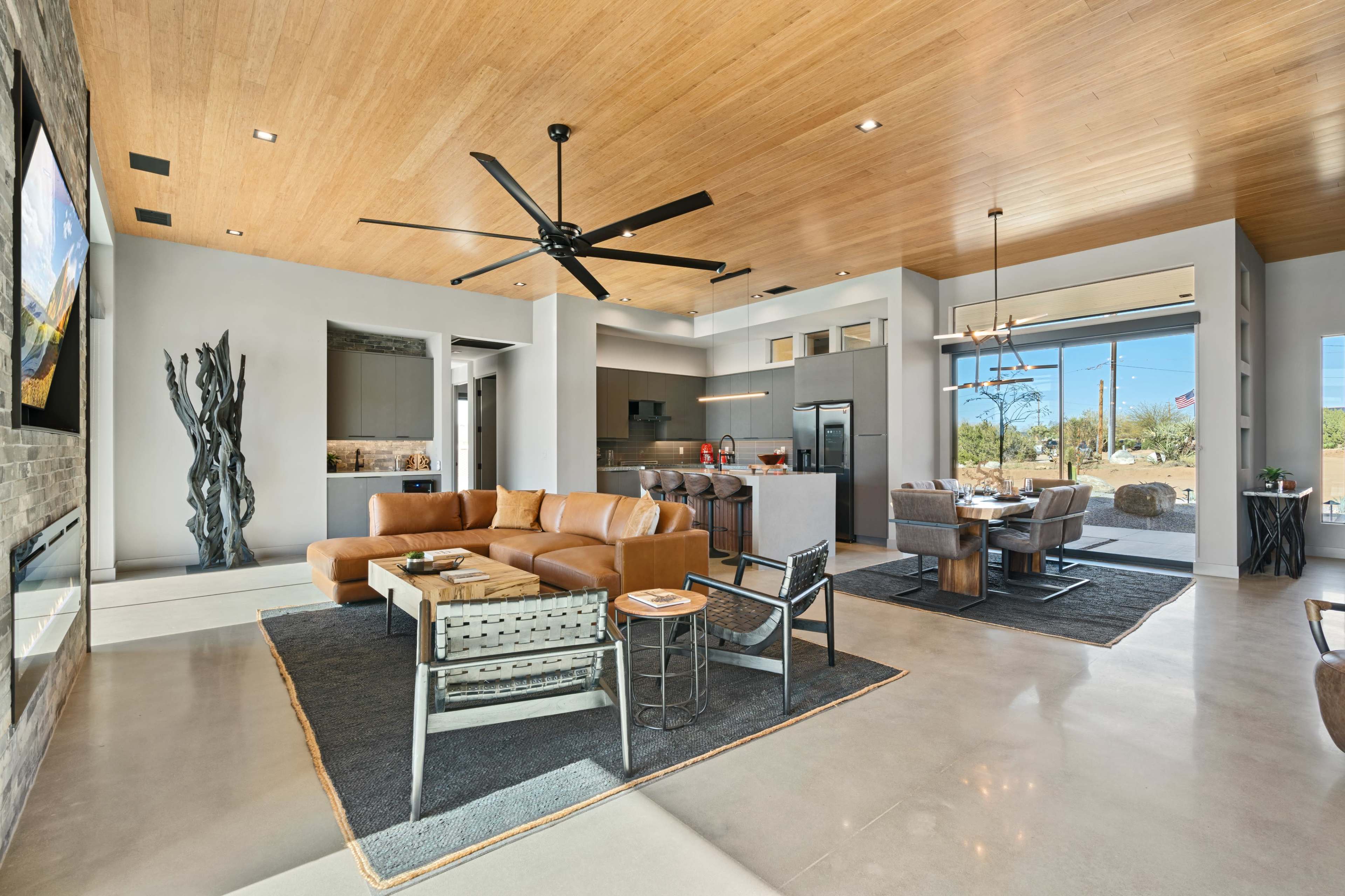 The image shows a modern living room with a large brown leather sofa, a dining area, and a kitchen, featuring wooden ceilings and large windows that offer outdoor views.
