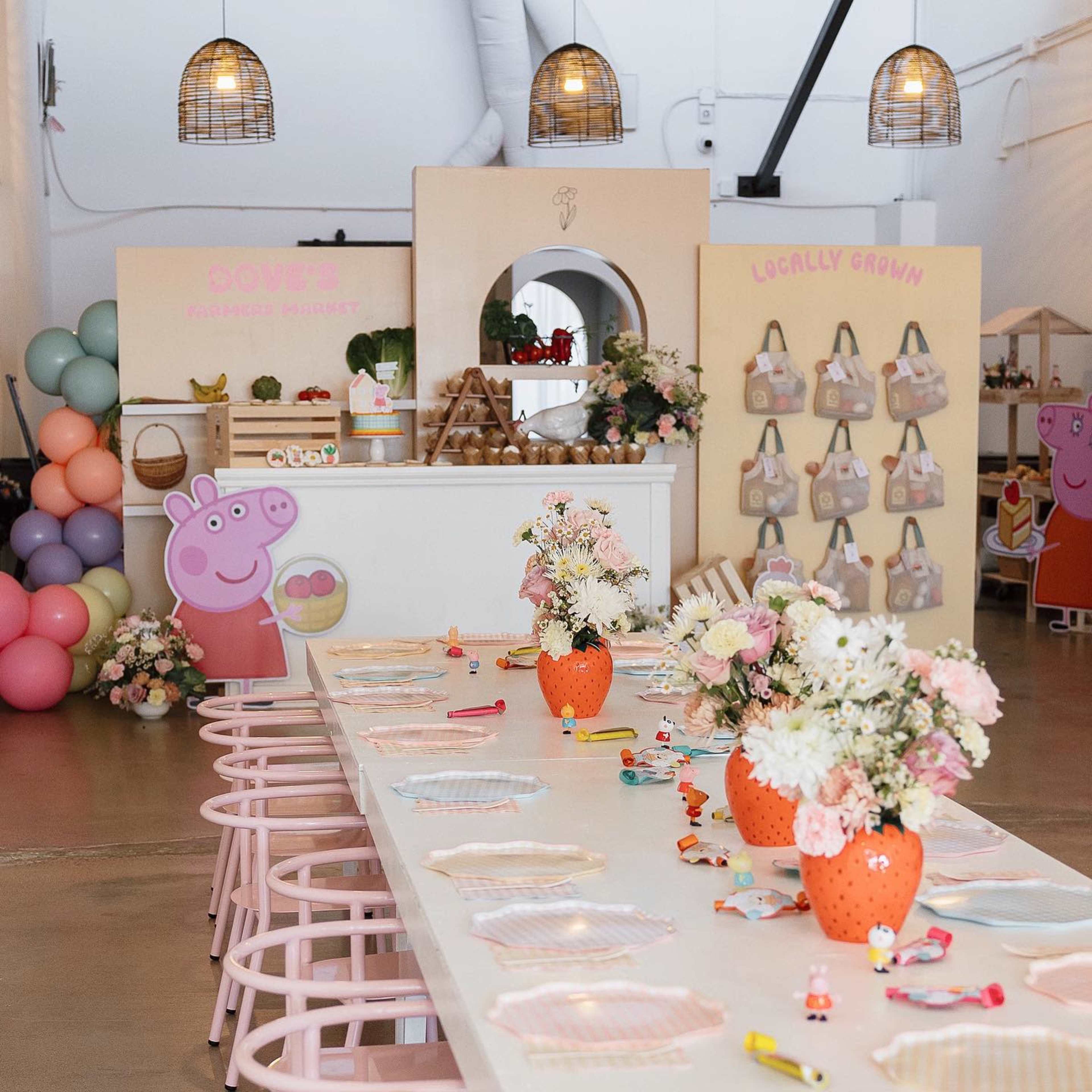 The image shows a decorated event space with a long table adorned with flowers, pastel-colored placemats, and party favors, while a Peppa Pig-themed backdrop and colorful balloon arrangements enhance the atmosphere.