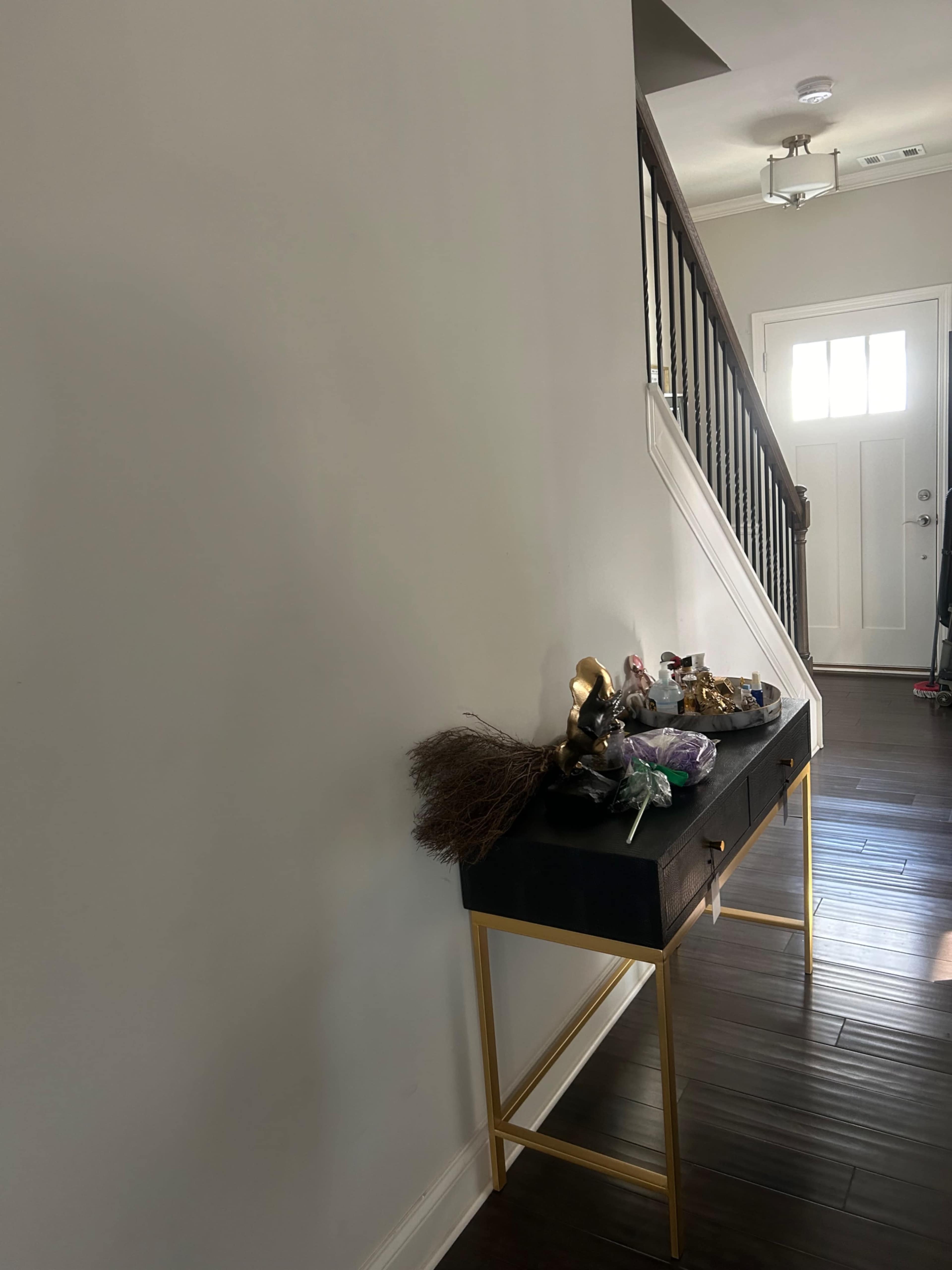 The image shows a hallway with a staircase on the right, featuring a narrow table on the left adorned with decorative items and a broom.