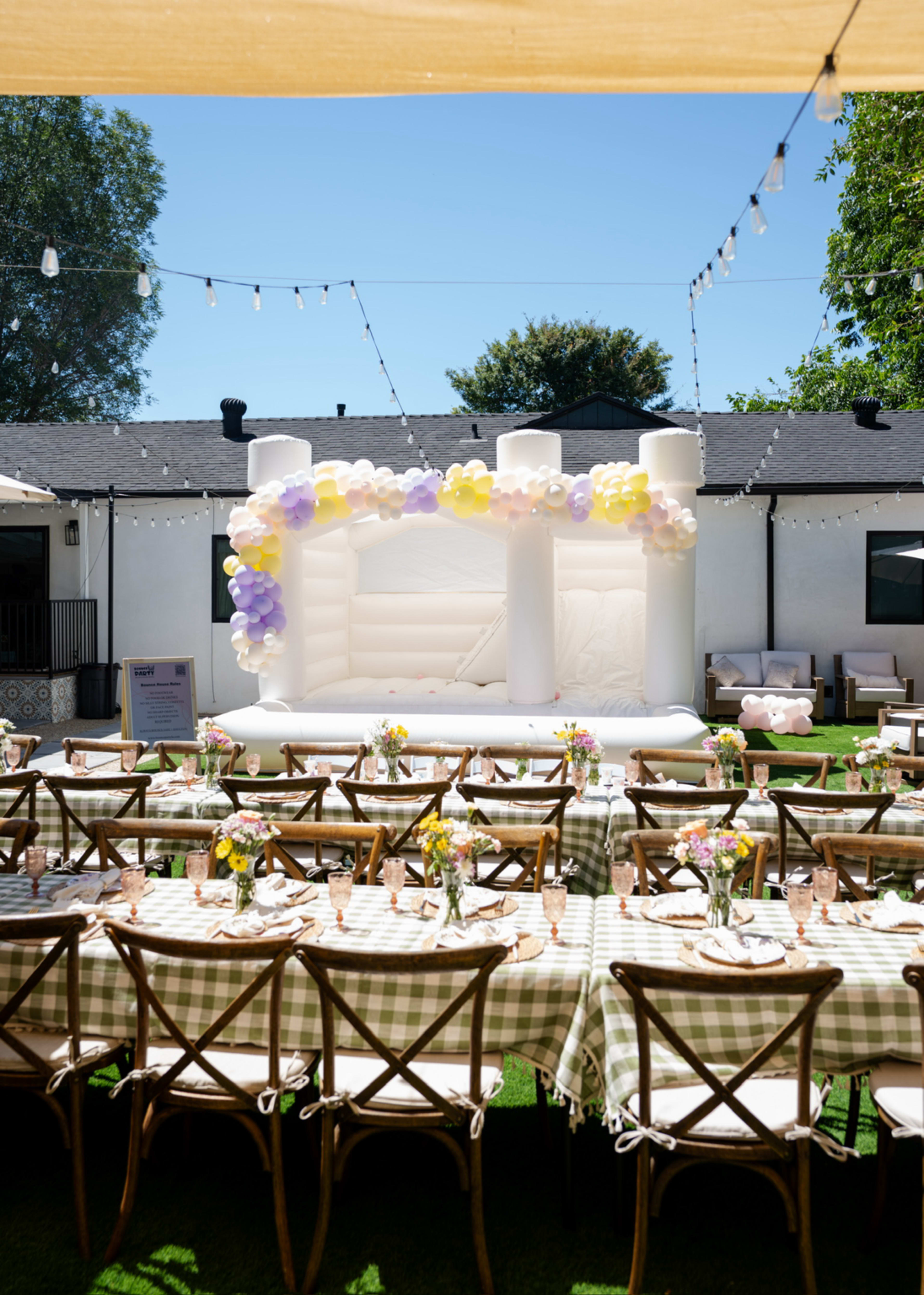 A spacious outdoor party setup features a long table adorned with checkered tablecloths, set with colorful flower centerpieces and glasses, alongside a large white inflatable structure decorated with balloons.