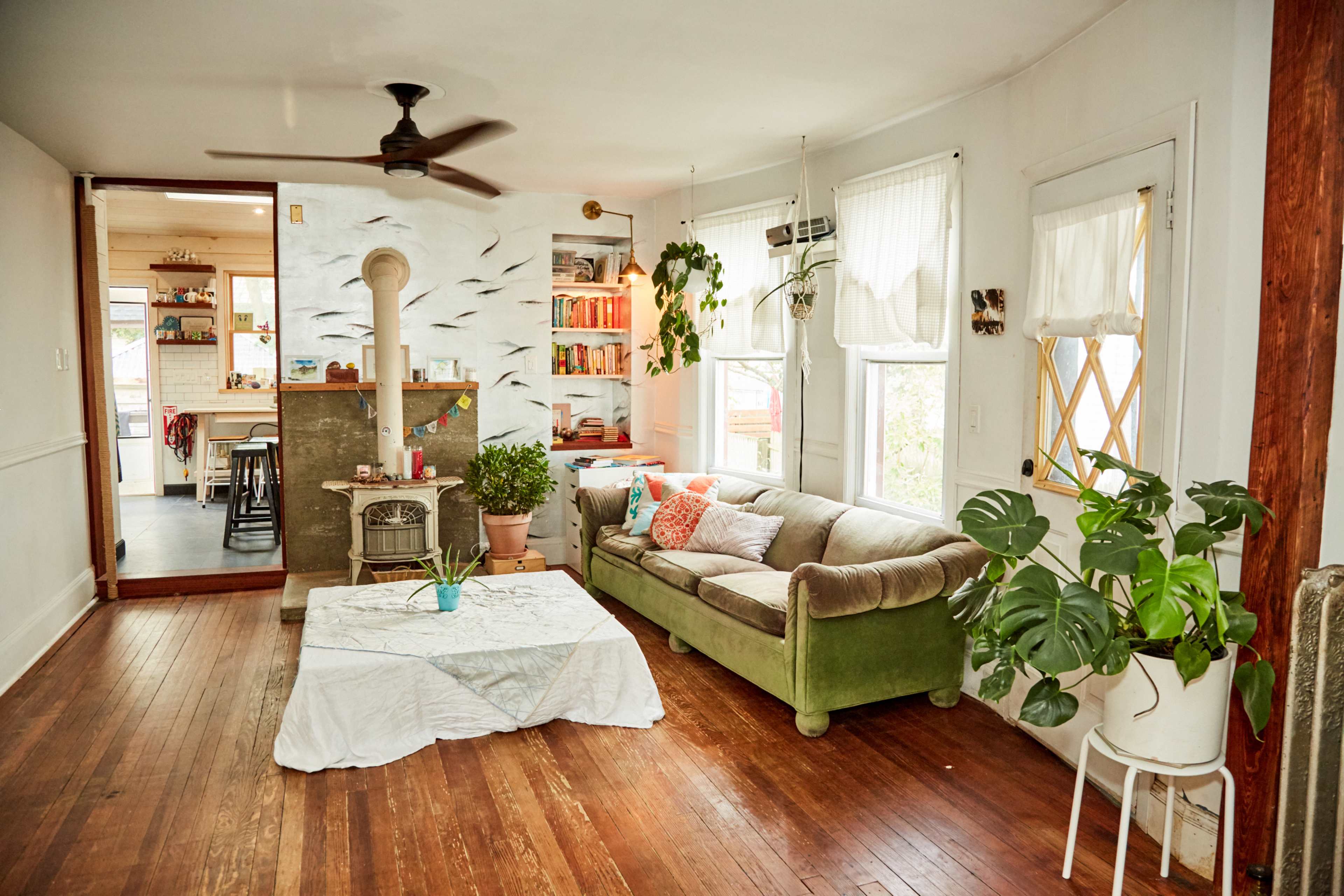 A cozy living room with a green sofa, wooden floor, and a wood stove, surrounded by plants and decorated with colorful cushions and a table in the center.