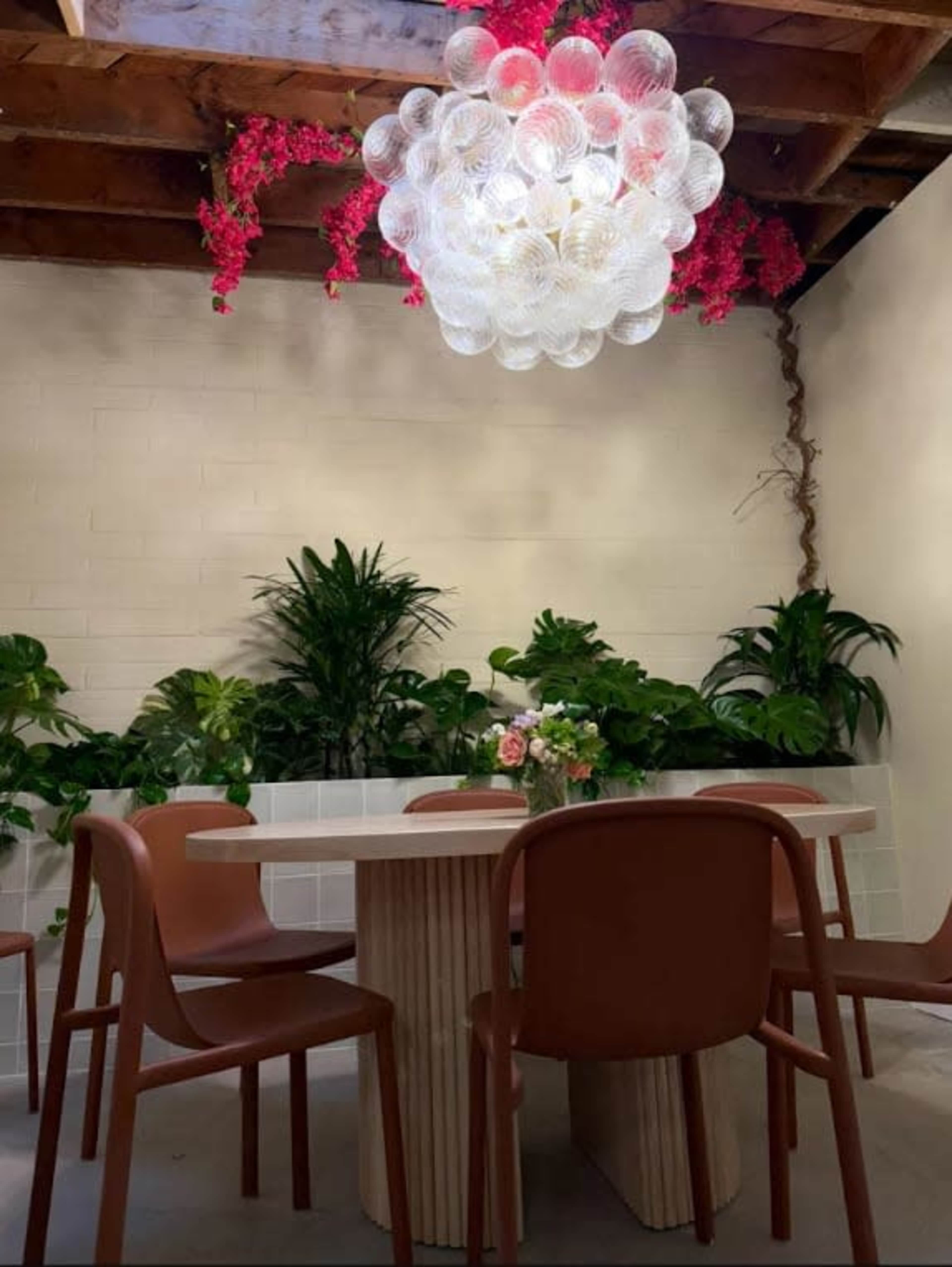 A dining area features a round table surrounded by brown chairs, illuminated by a decorative light fixture and adorned with various plants and flowers.