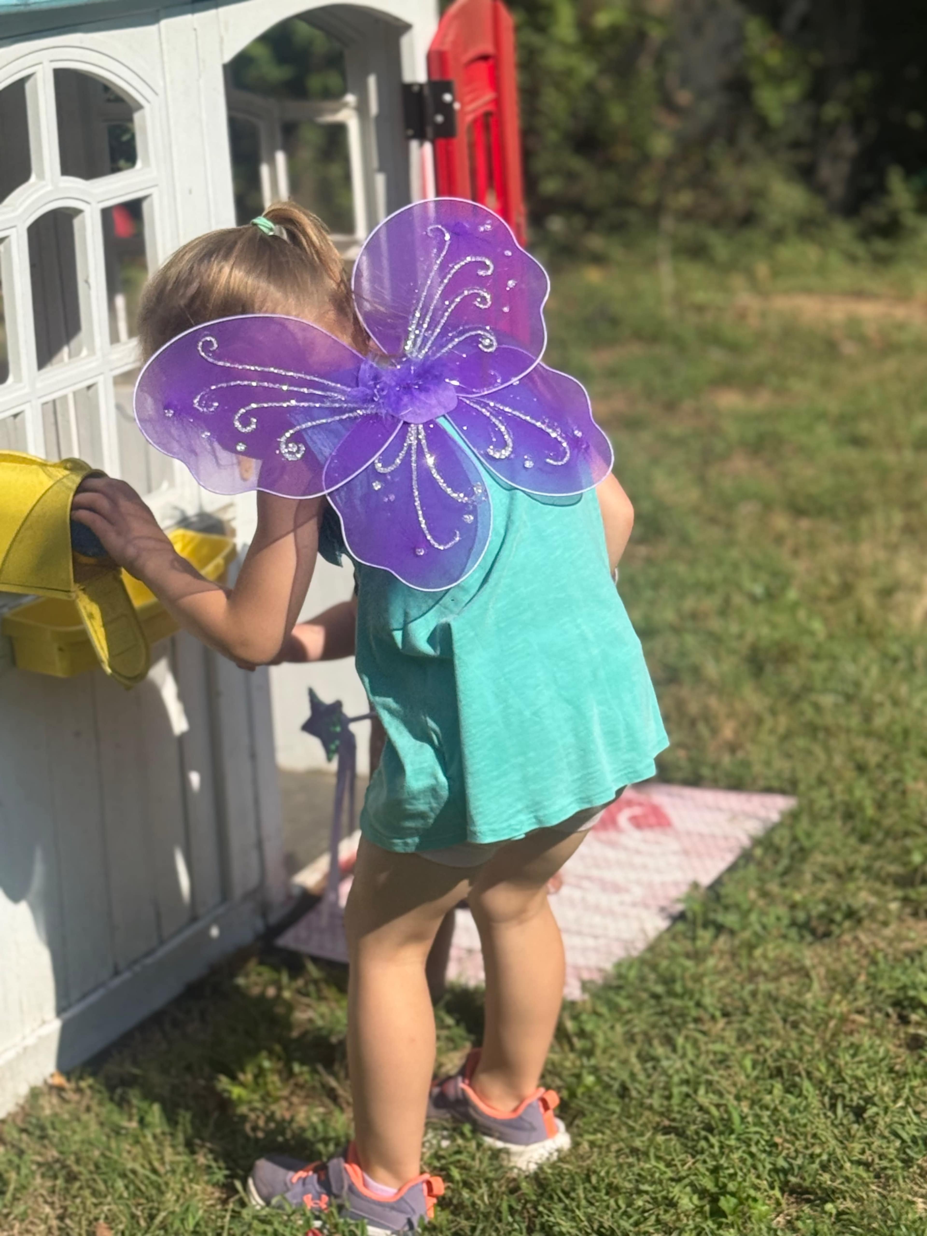 A young child wearing butterfly wings looks into a yellow mailbox outside a small playhouse.