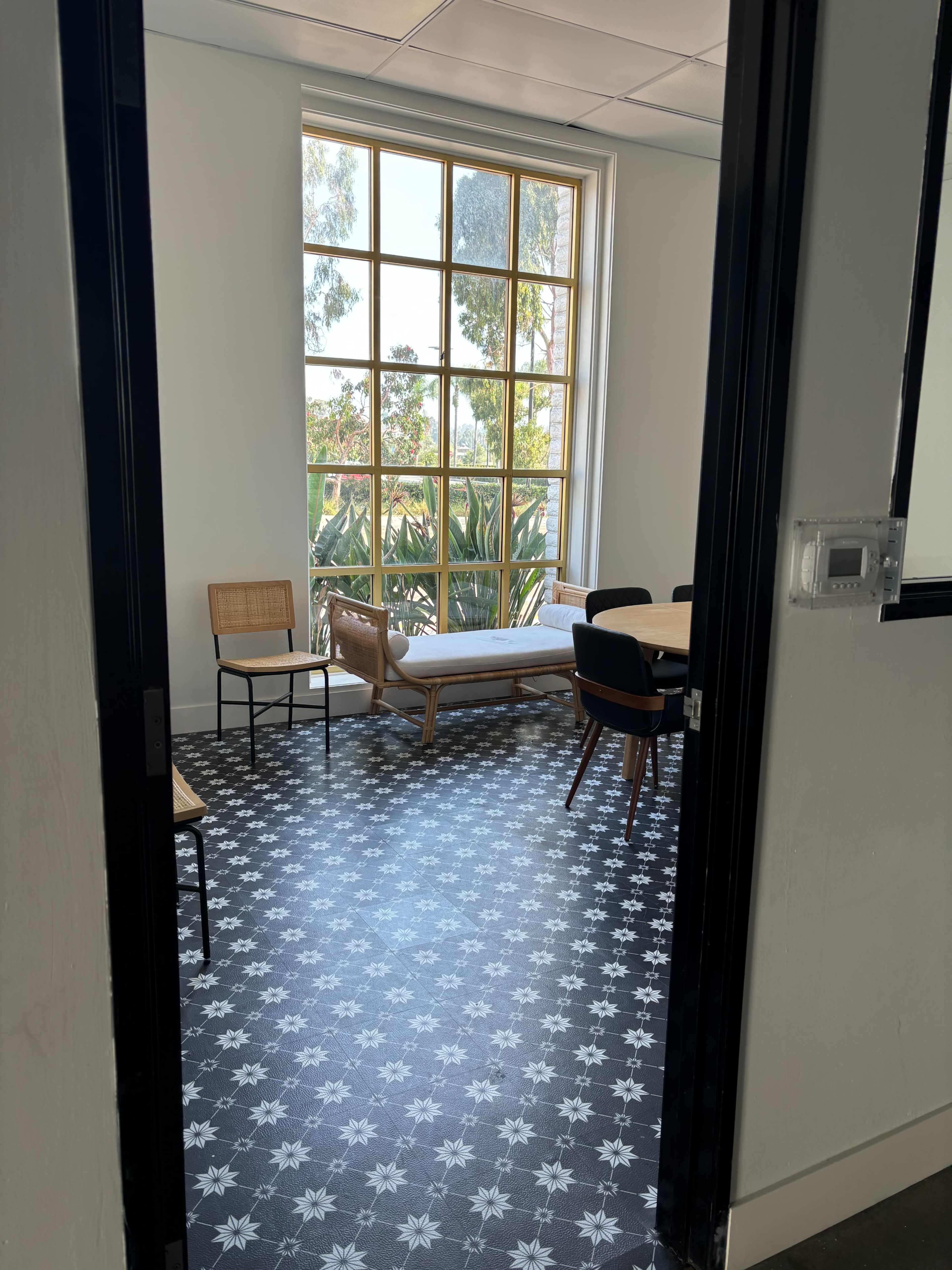 The image shows a room with a patterned floor, a large window, a seating area with a couch and chairs, and visible greenery outside.