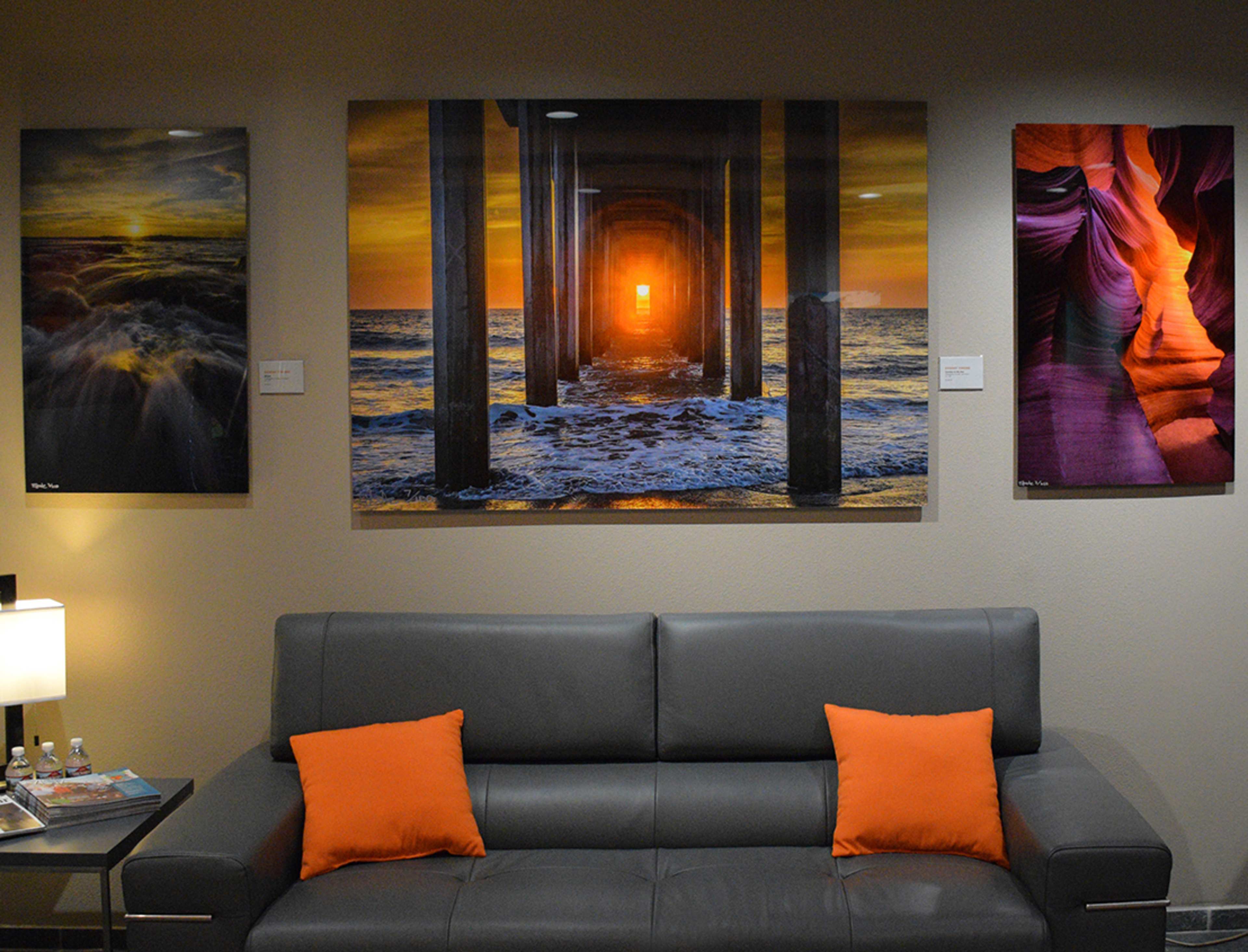 A gray sofa with orange cushions placed against a wall featuring three large art prints, including a sunset over the ocean viewed through pilings and an abstract canyon scene.