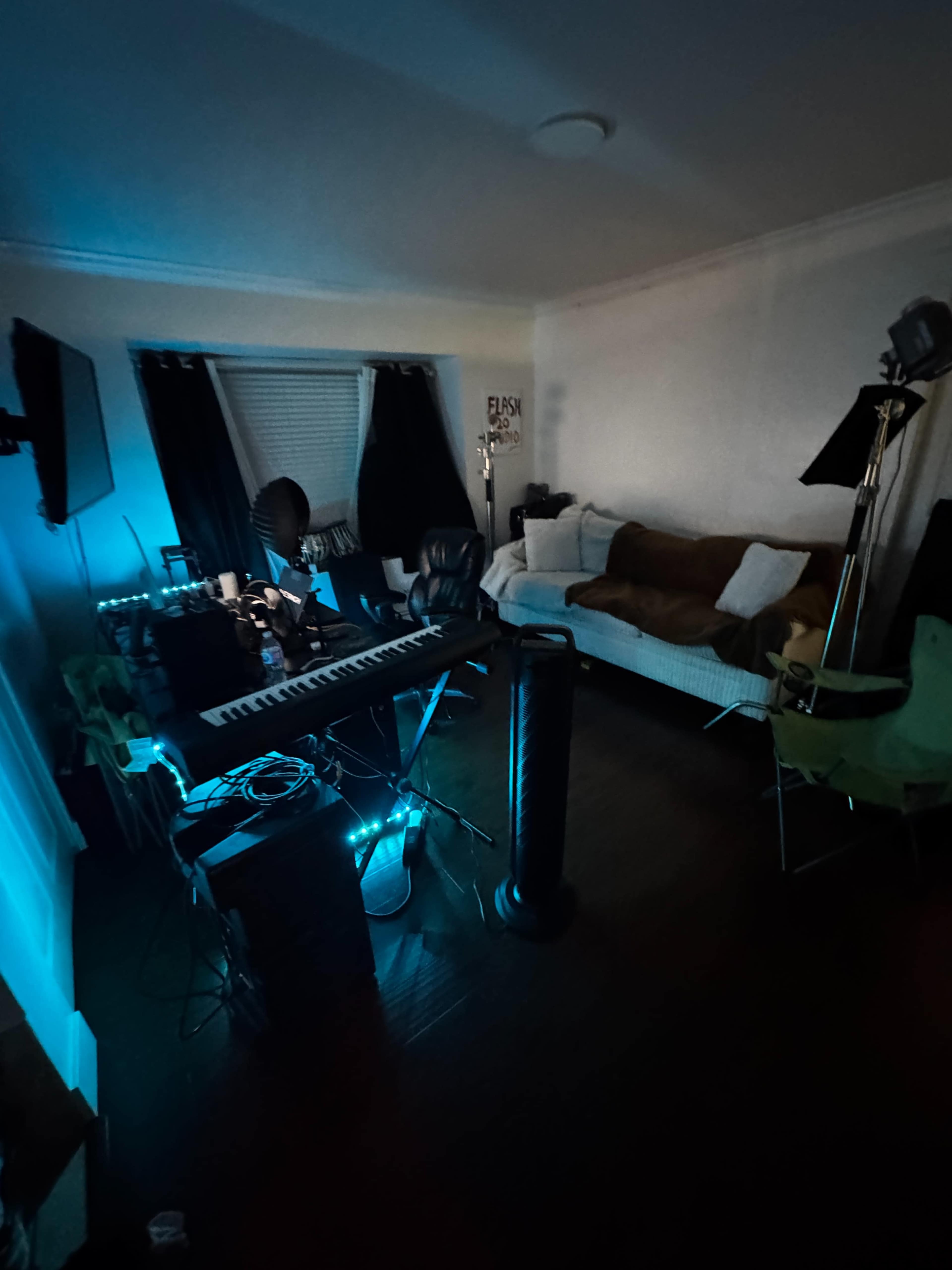 The photo shows a dimly lit room featuring a keyboard, a fan, and a couch with blankets, along with some chairs and various equipment scattered around.