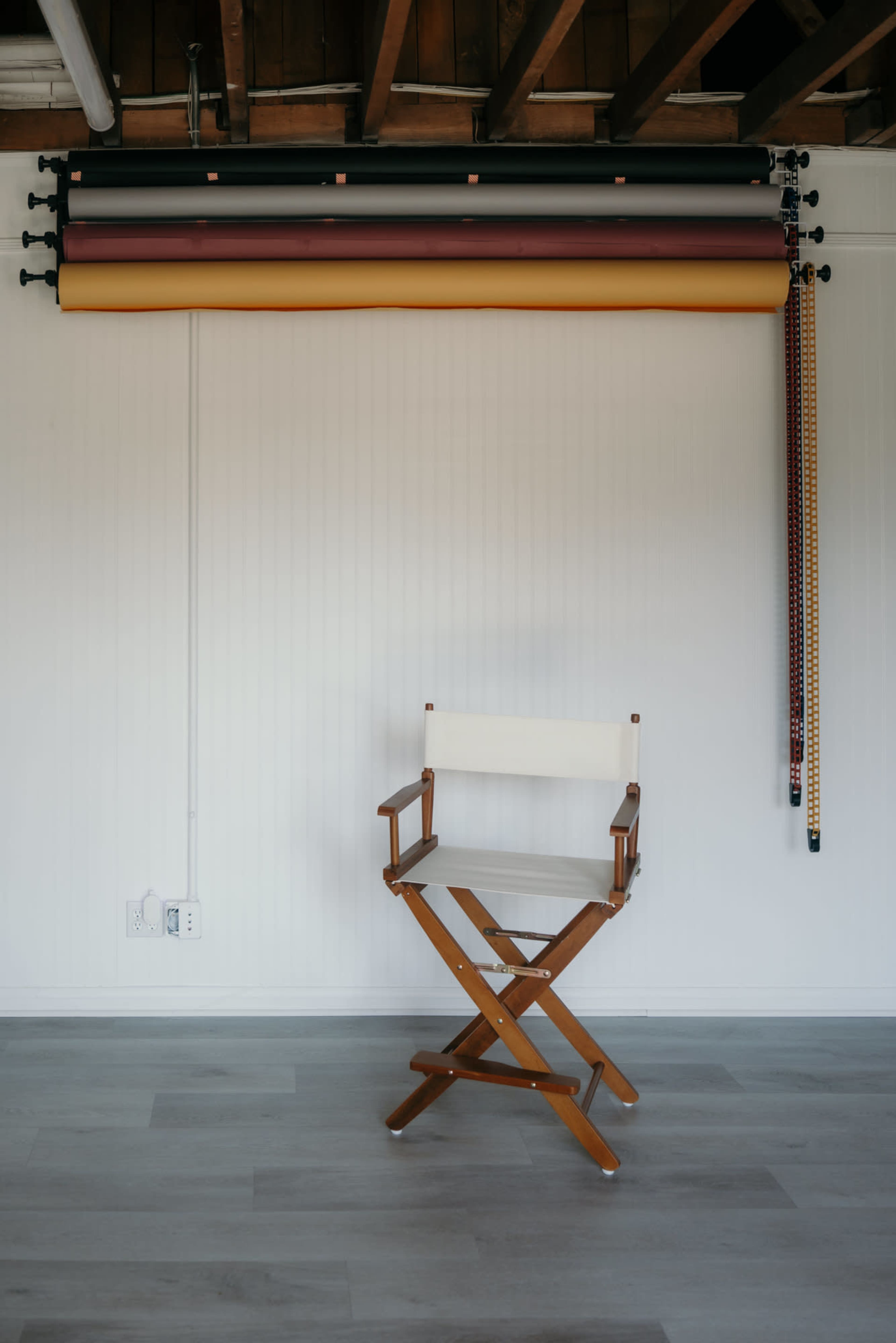 The image shows a wooden director's chair in front of a wall with rolled-up backdrop materials above it.