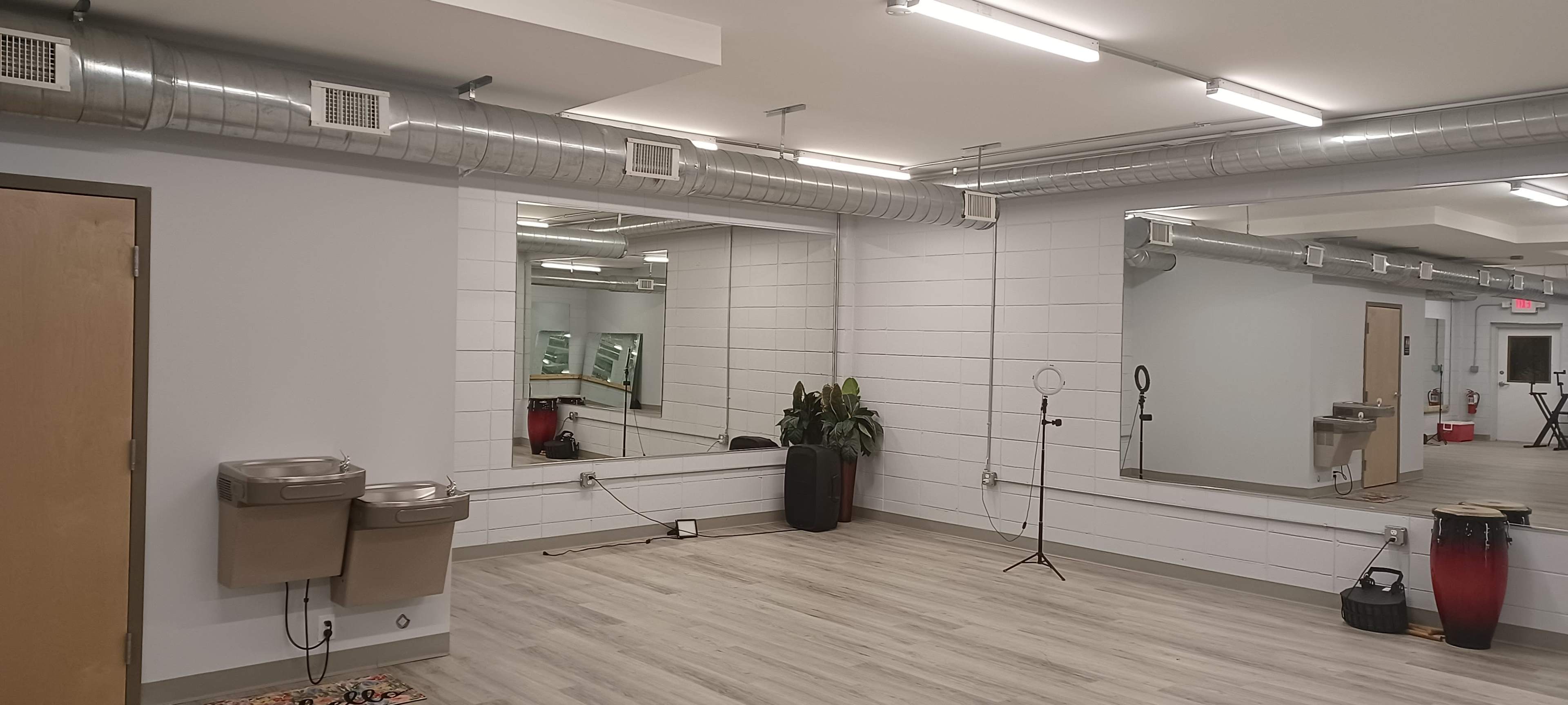 The image shows a spacious room with large mirrors, metal ventilation ducts on the ceiling, and some fixtures like a plant and two water fountains.