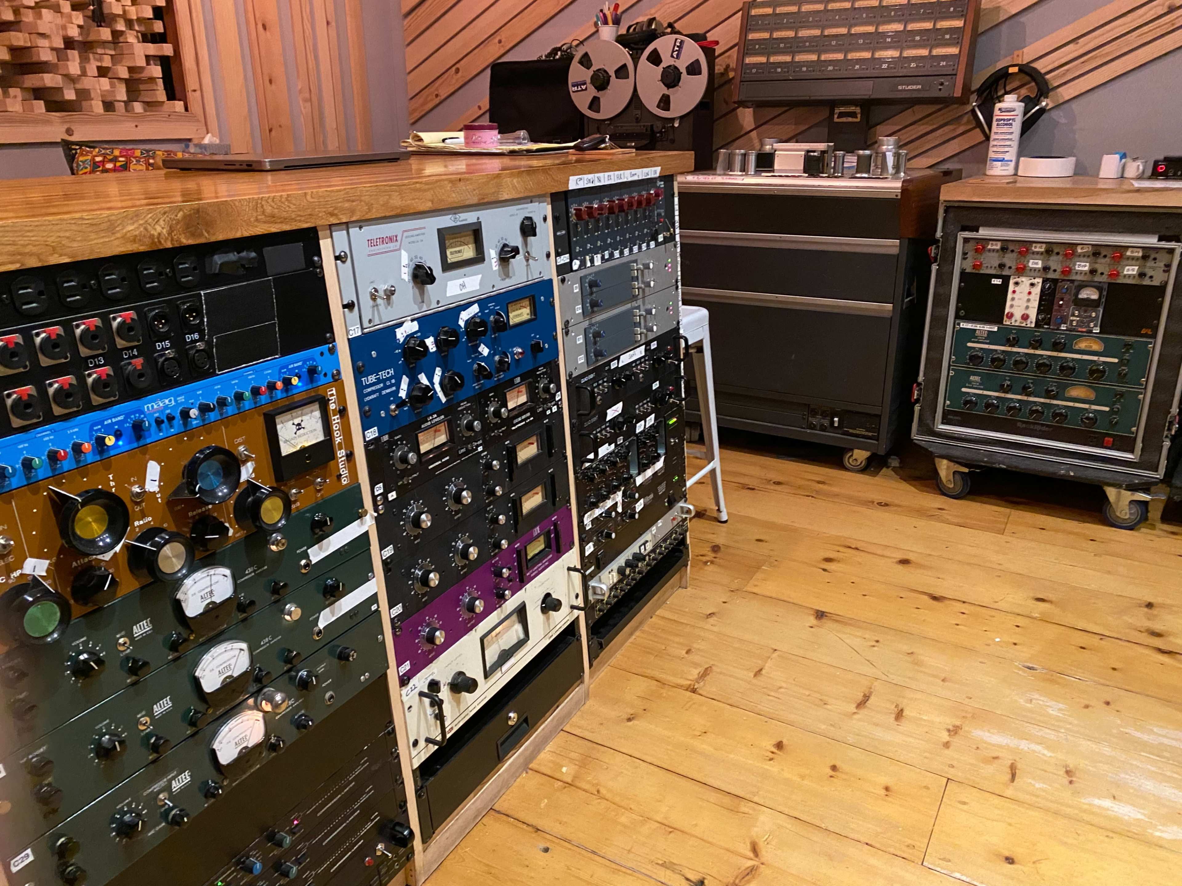 A recording studio with various audio equipment, including a console, tape reels, and rack-mounted gear, arranged against wooden walls and a wooden floor.