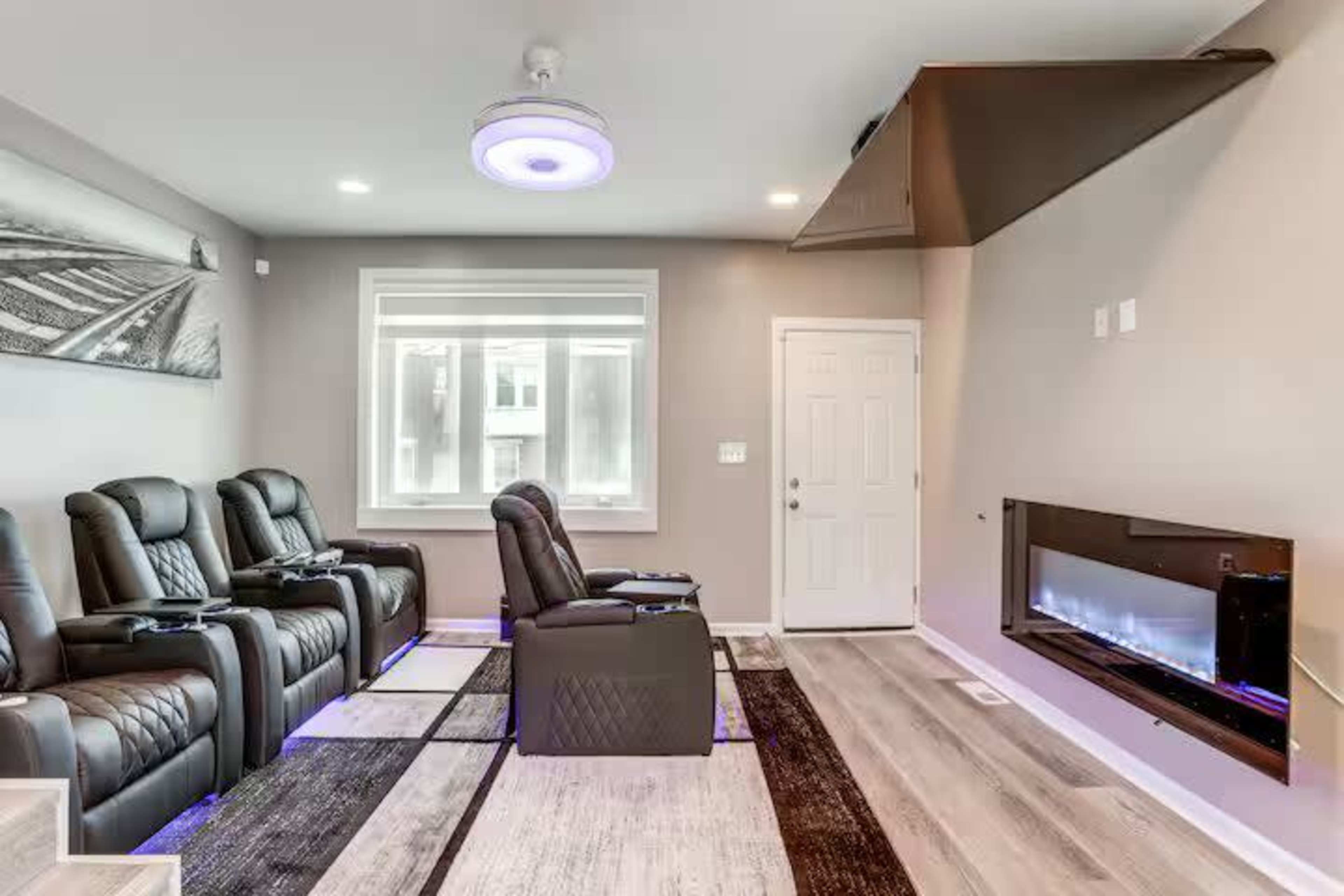 The image shows a modern home theater with reclining seats, a wall-mounted electric fireplace, and large windows allowing natural light.