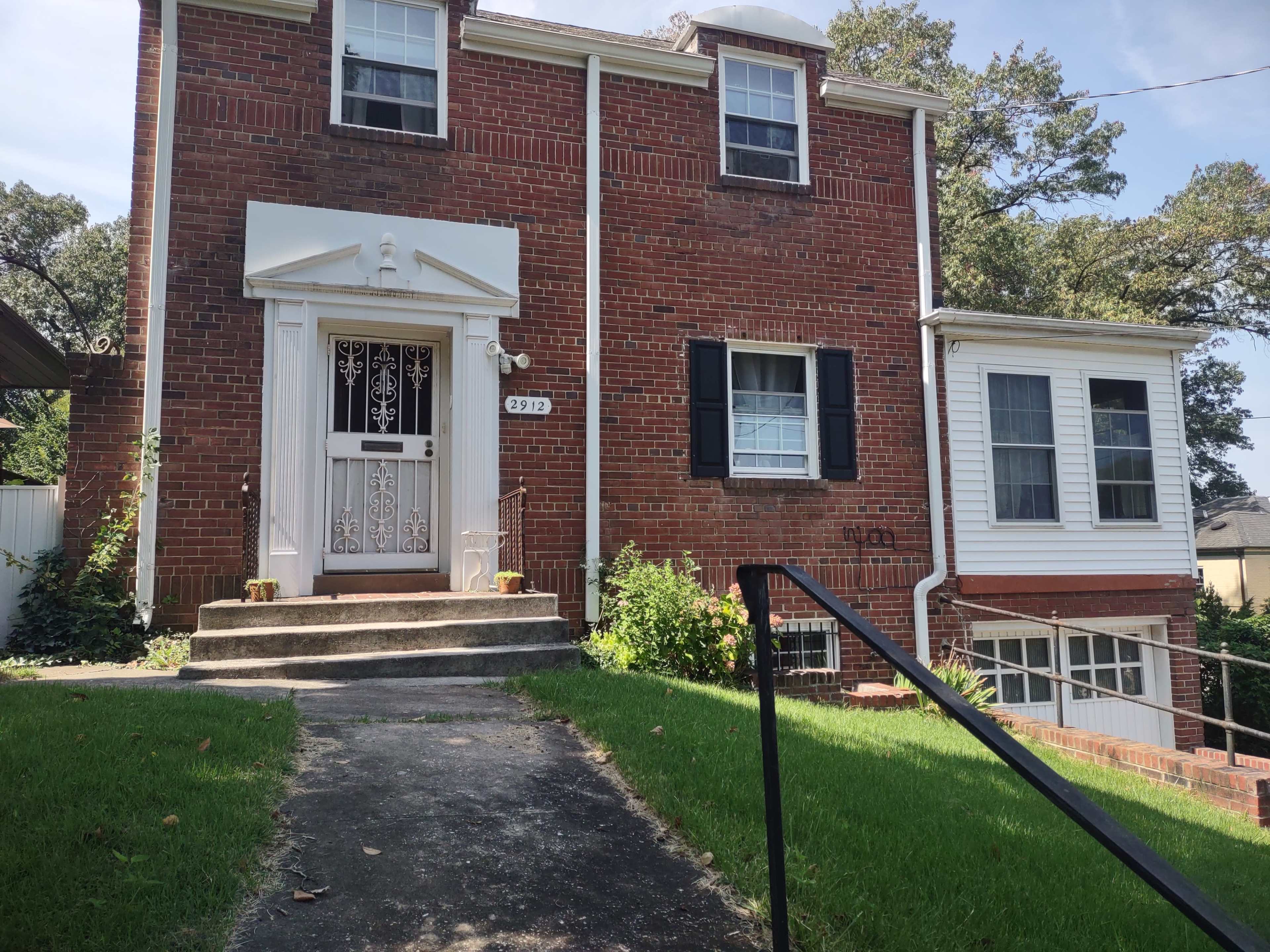 A two-story brick house with white trim, a front porch, and a pathway leading to the entrance.