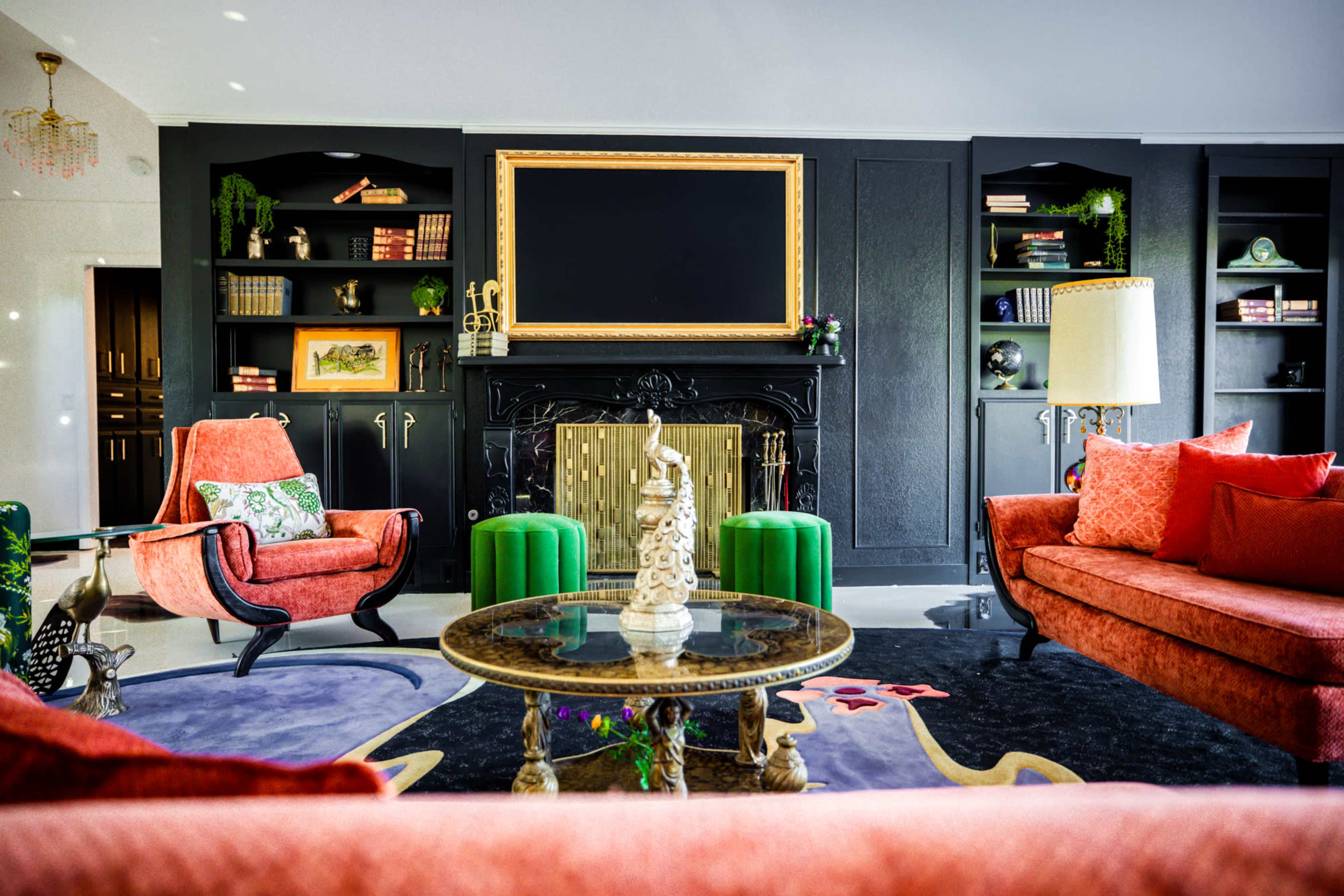 The image shows a stylish living room with two orange sofas, green side tables, a round coffee table, and built-in black shelves filled with decor and books.