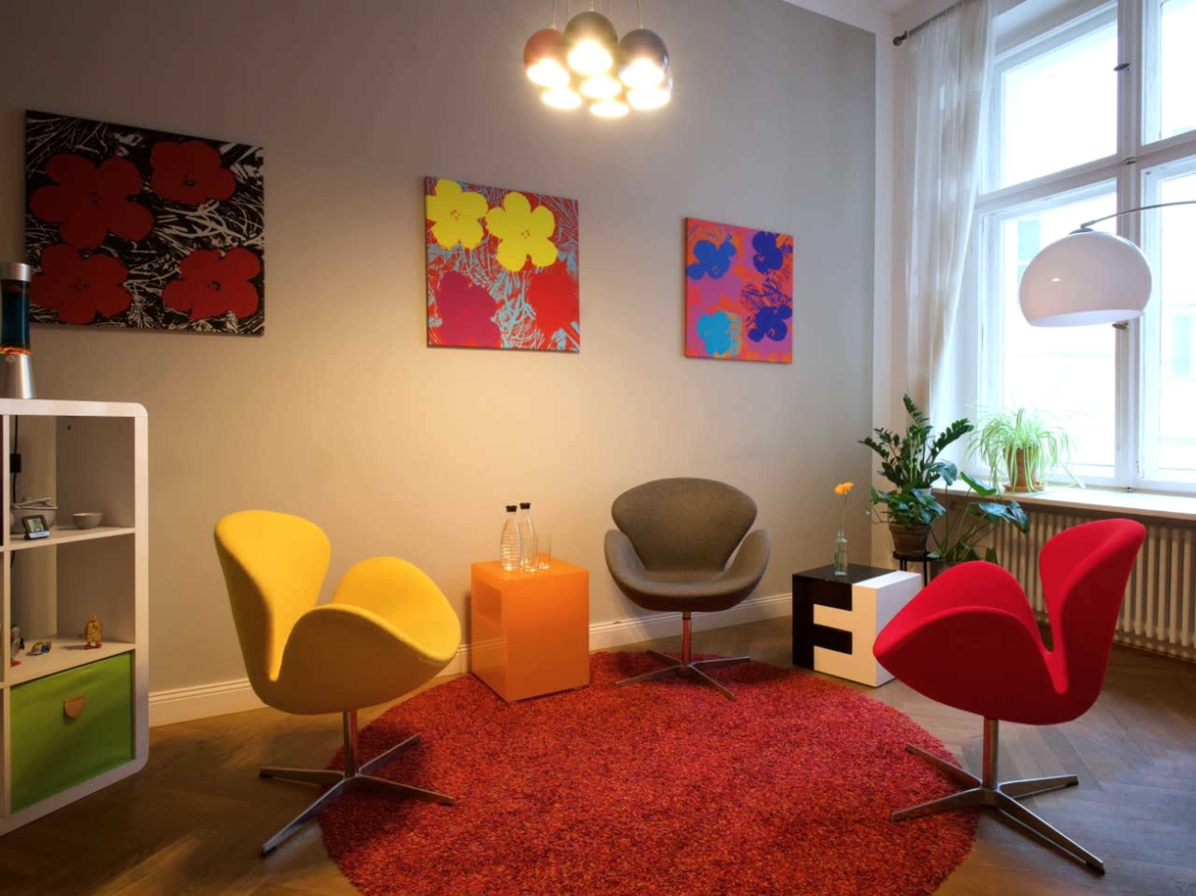 A modern seating area with three colorful chairs arranged on a circular red rug, surrounded by framed artworks on the walls and natural light coming through a window.