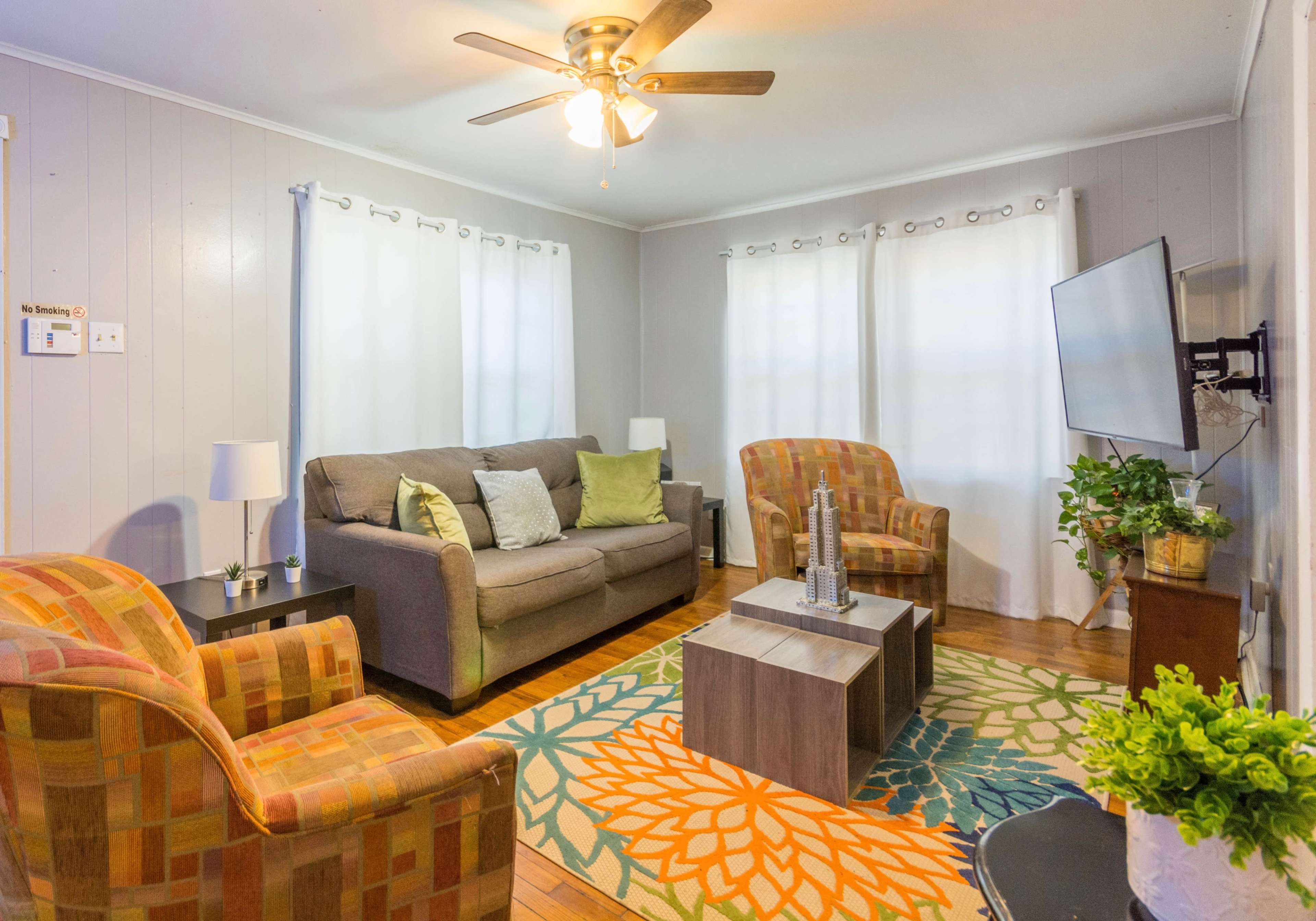 A cozy living room featuring a gray sofa, two patterned armchairs, a coffee table, and a television mounted on the wall, with light-colored curtains framing the windows.