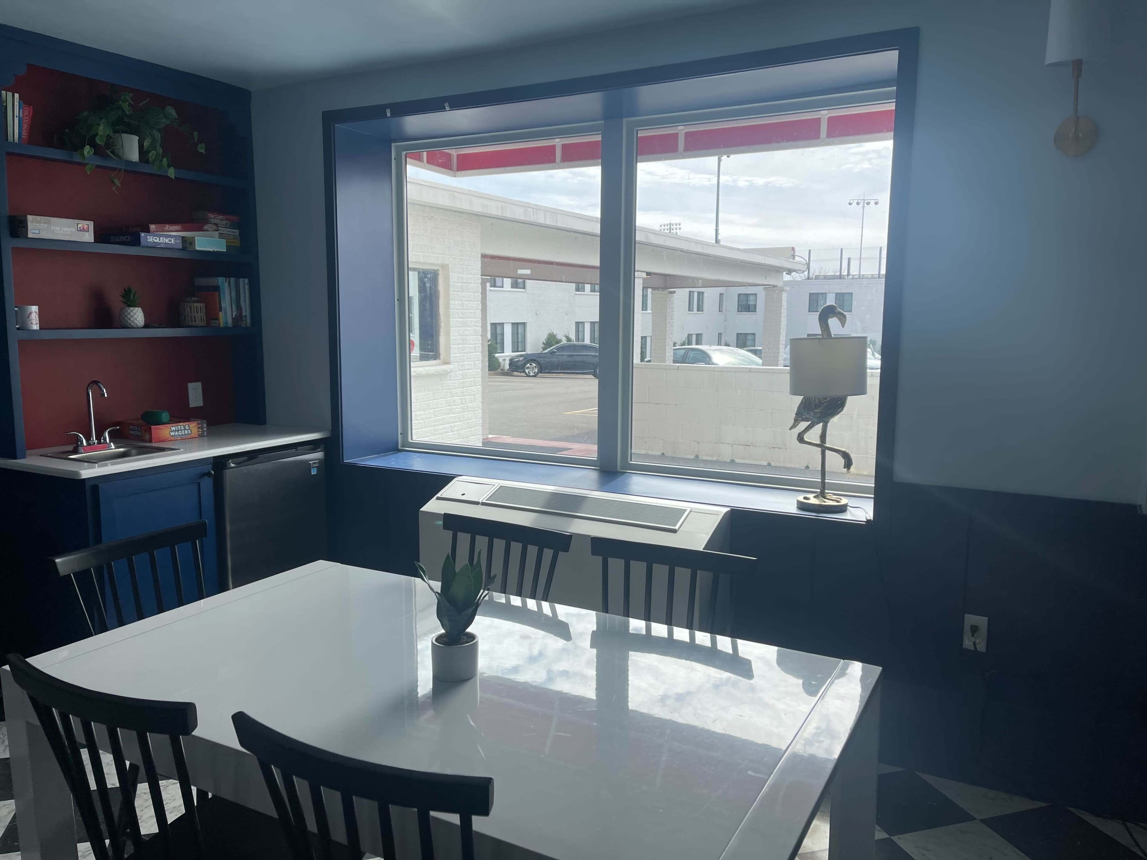 A brightly lit kitchen features a table with chairs, a small plant centerpiece, and a window overlooking an exterior view with parked cars.