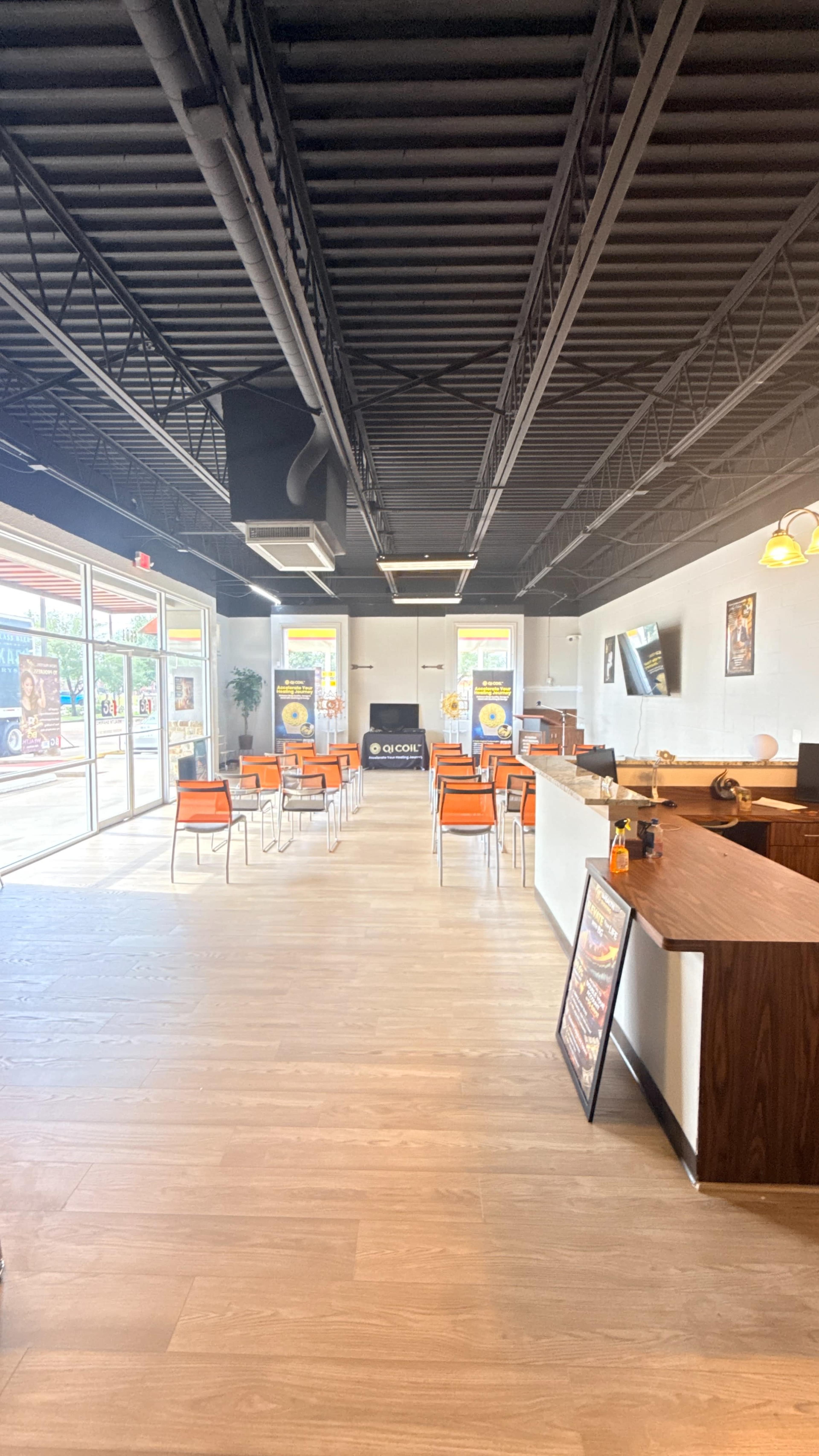 The image shows a modern, spacious interior of a restaurant with orange chairs arranged at tables and large windows allowing natural light to enter.