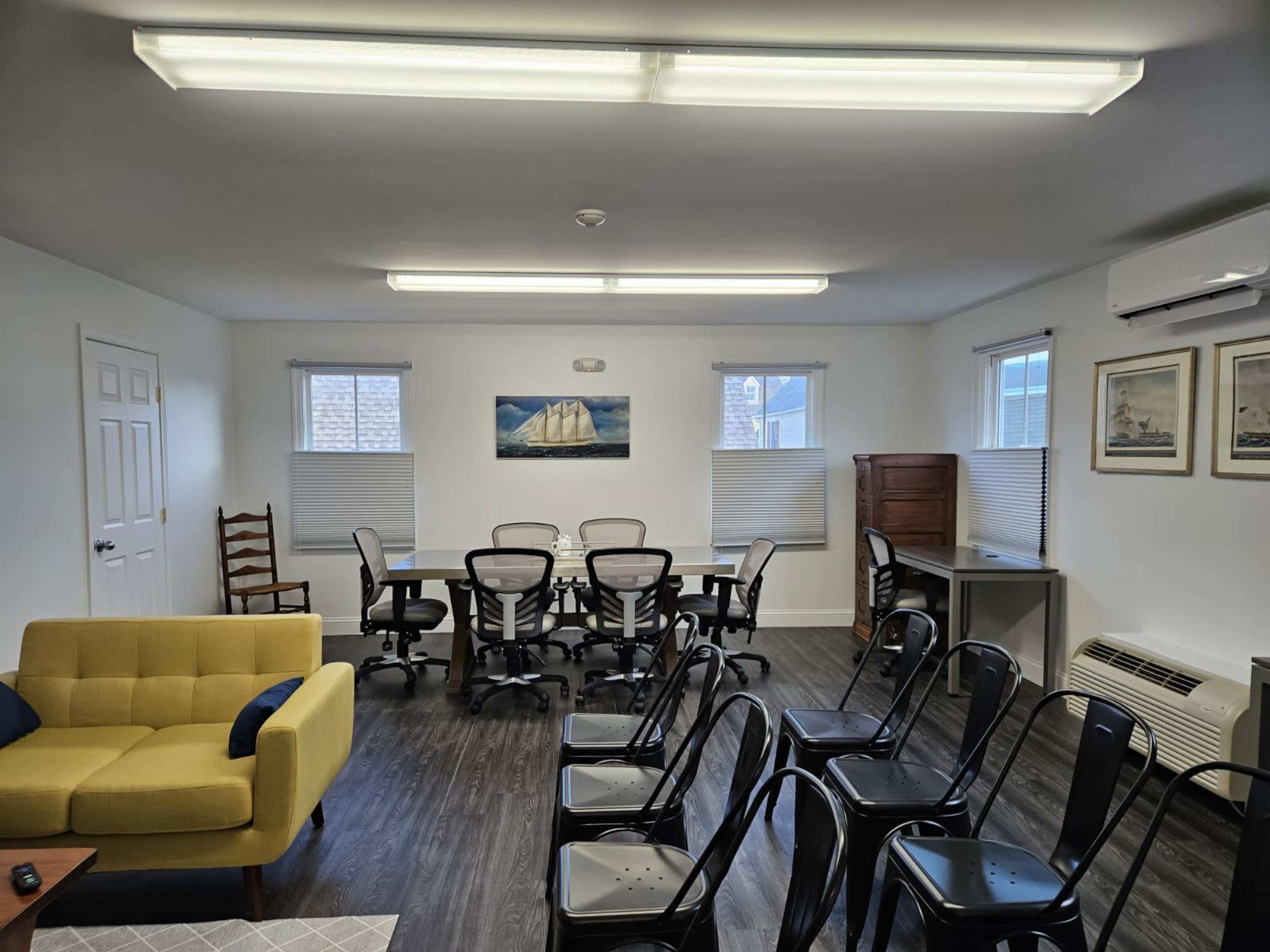 The image shows a well-lit meeting room with a large table surrounded by rolling chairs, a yellow sofa in the corner, and framed nautical artwork on the walls.