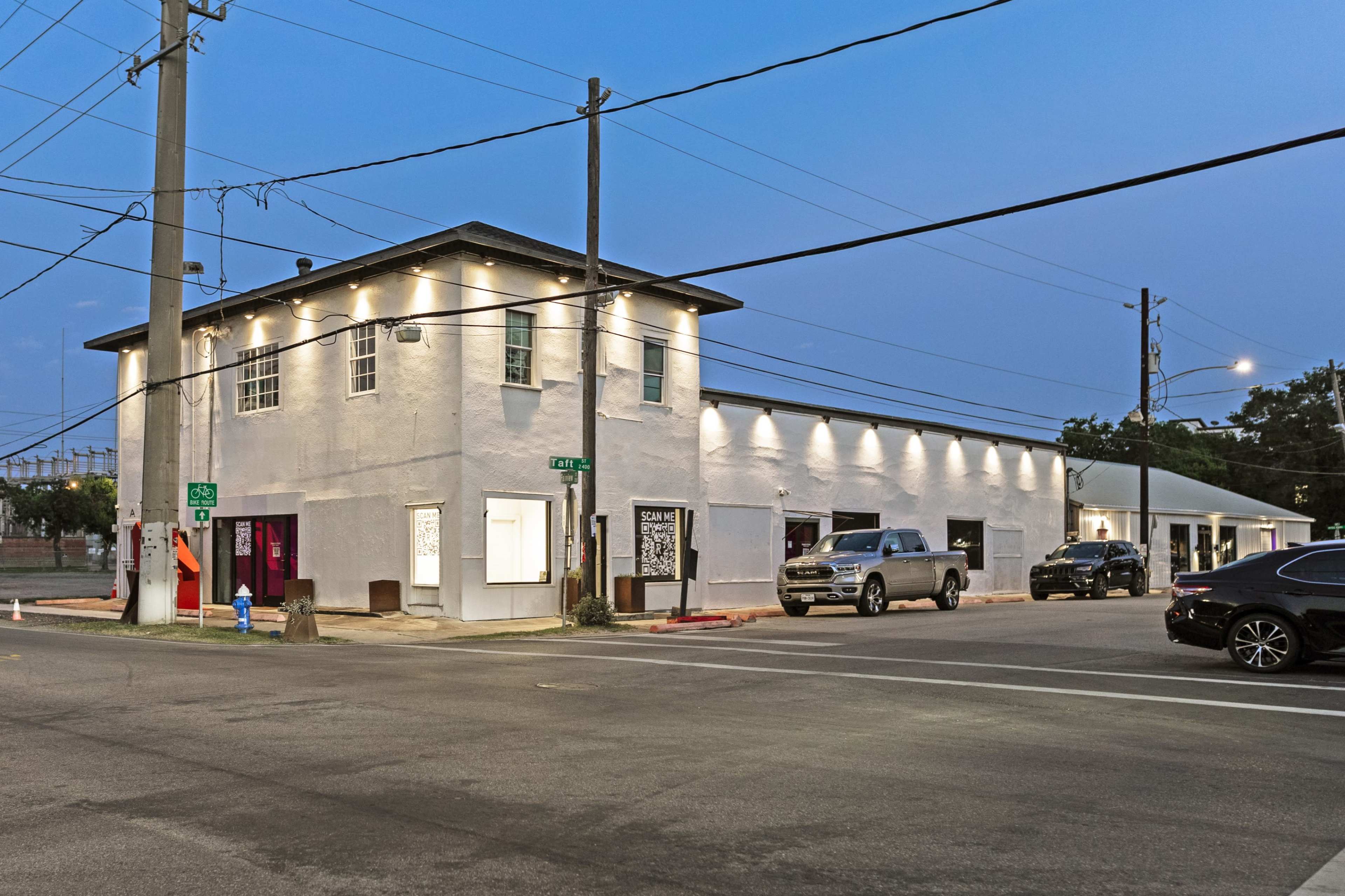 A two-story white building with illuminated windows sits at the corner of a street intersecting with a paved road, flanked by parked cars and utility poles.