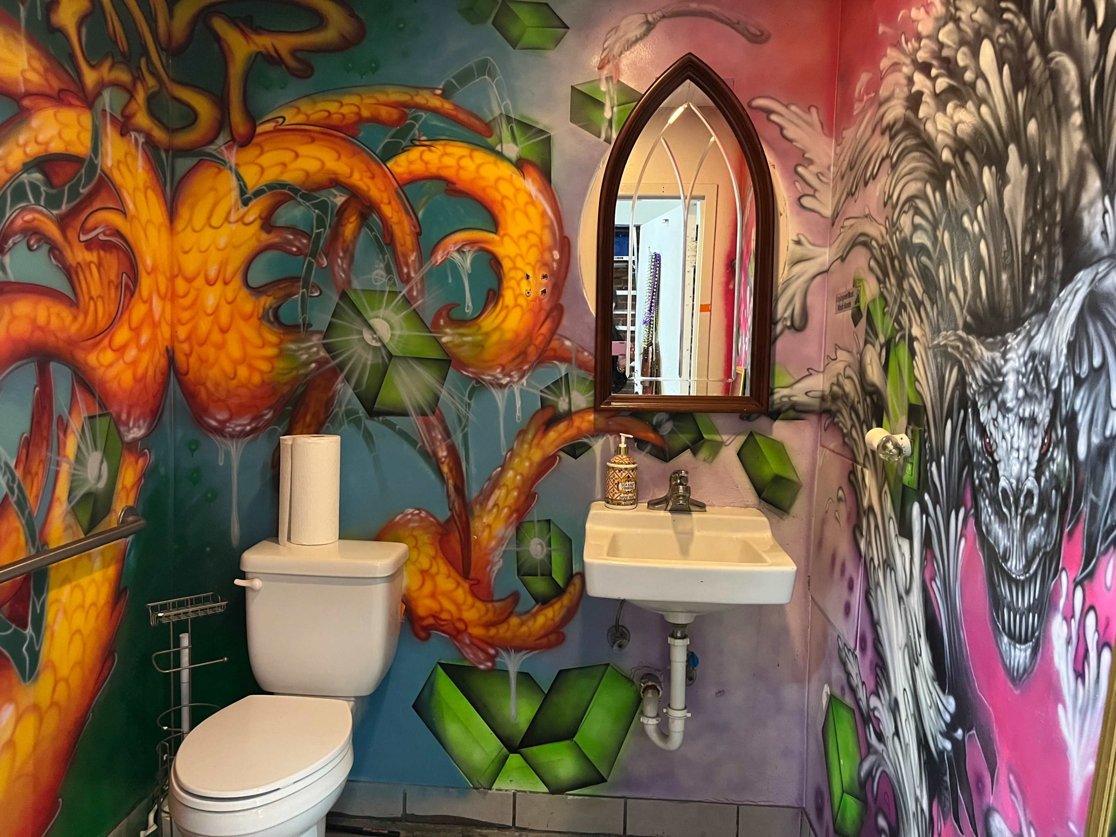 A small bathroom with vibrant, colorful graffiti covering the walls, featuring a toilet, a sink, and a decorative mirror.