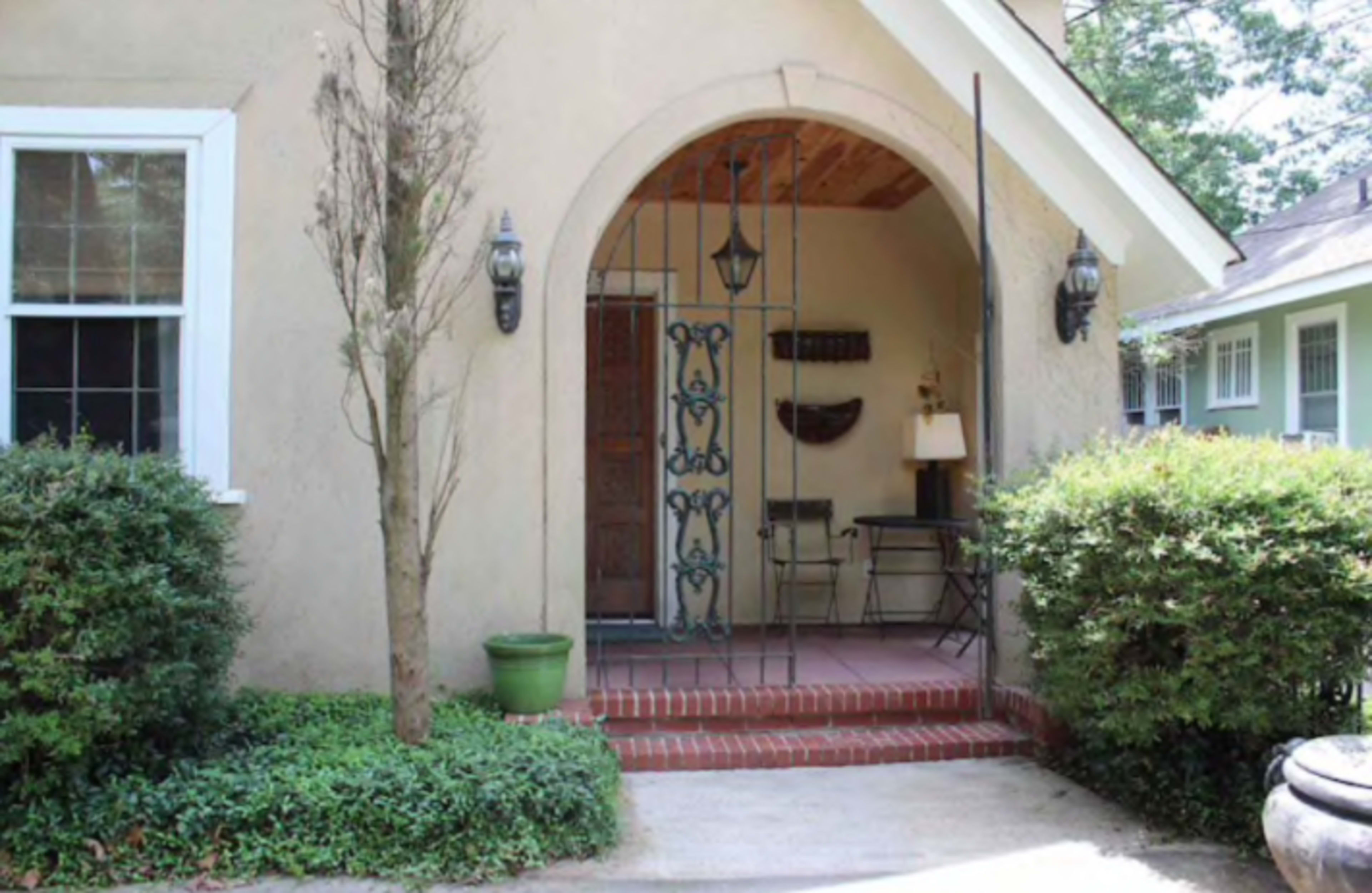 A small entrance with an arched doorway, featuring a wrought iron gate, brick steps, potted plants, and a table with a lamp inside.