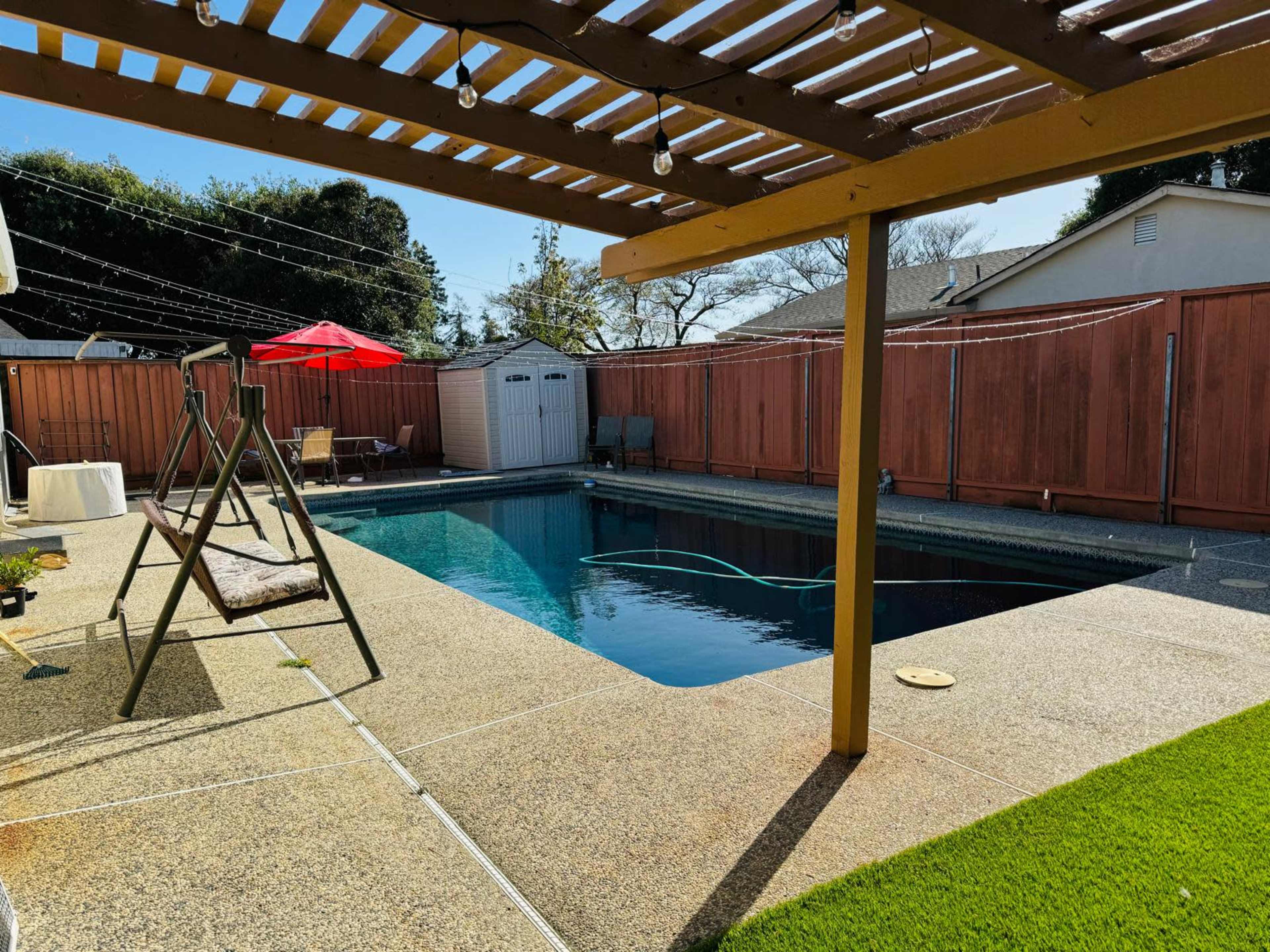 A backyard scene features a swimming pool surrounded by a concrete deck, a swing set, and a patio area with an umbrella and lawn.