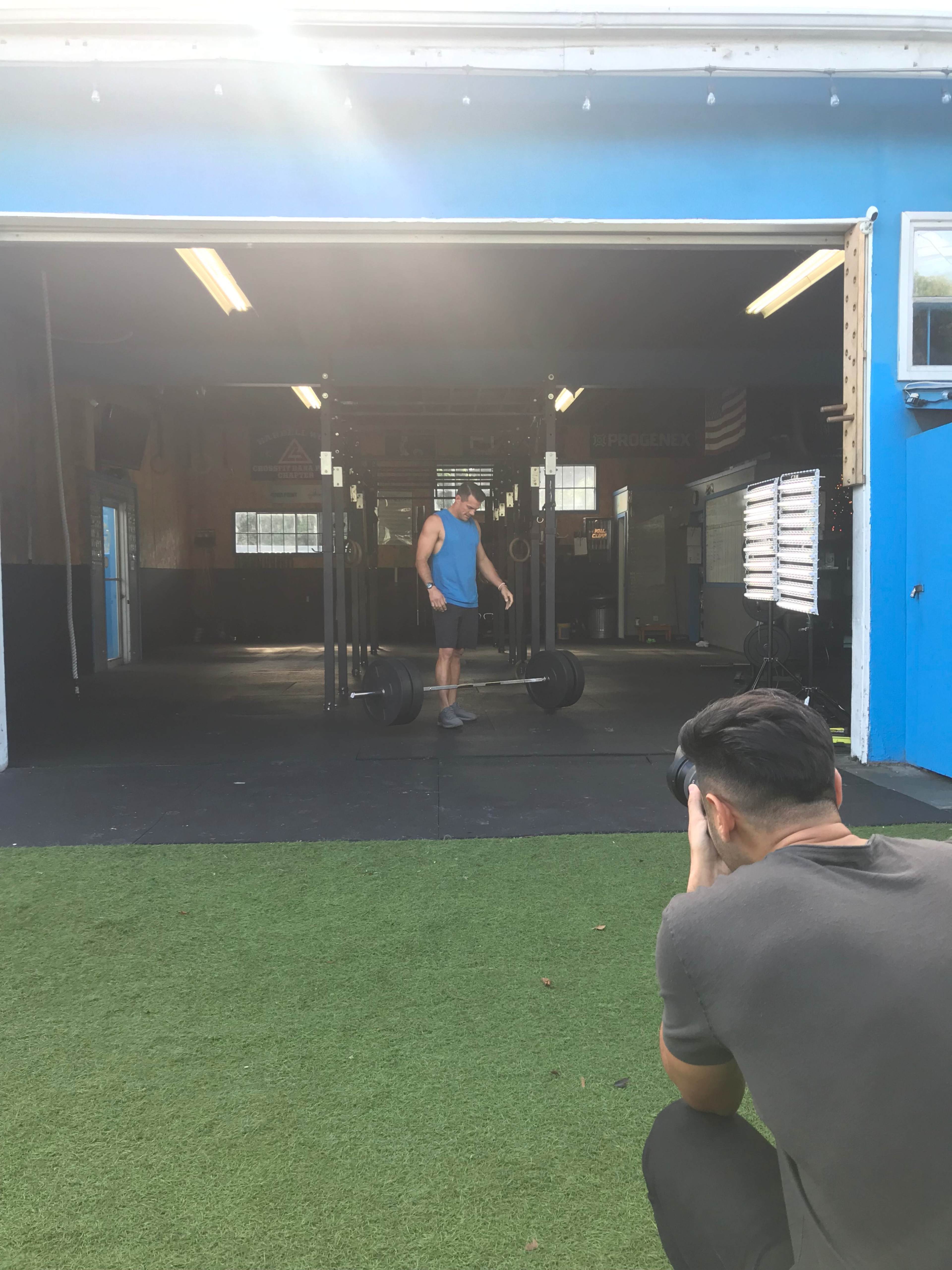 A man prepares to lift a barbell in a gym while another person photographs him from a distance.