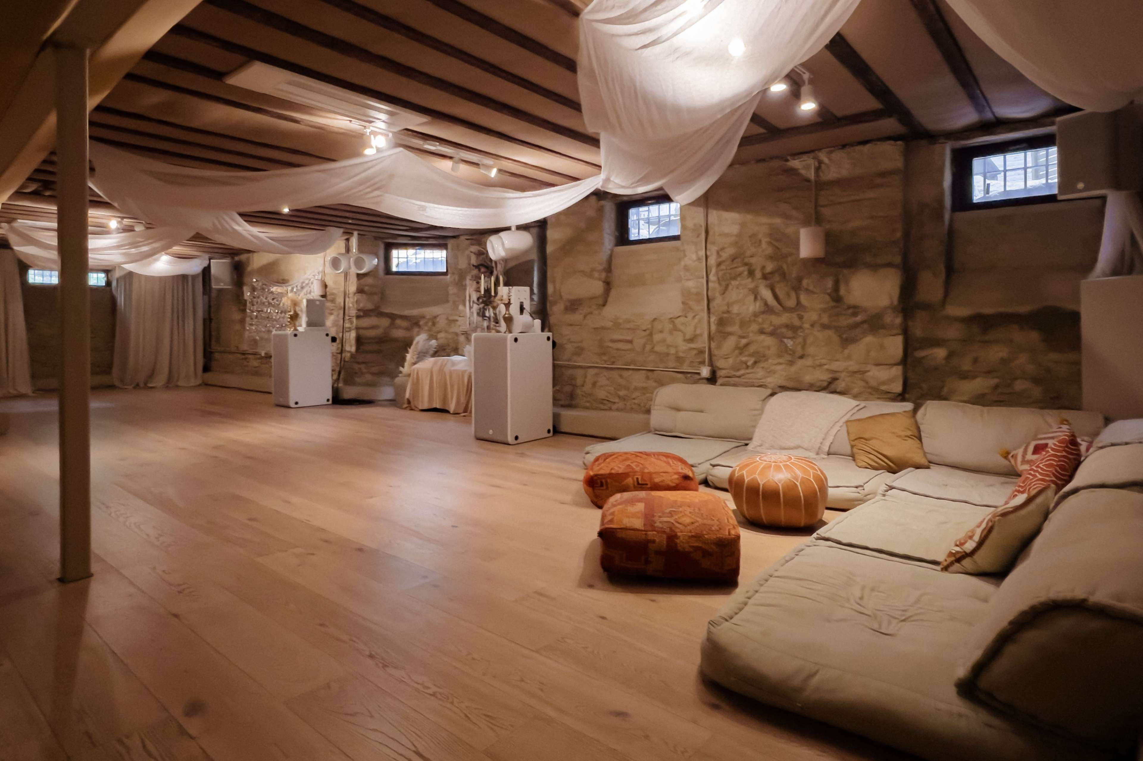 The image shows a spacious basement room with stone walls, soft lighting, a wooden floor, and several low seating arrangements adorned with cushions.