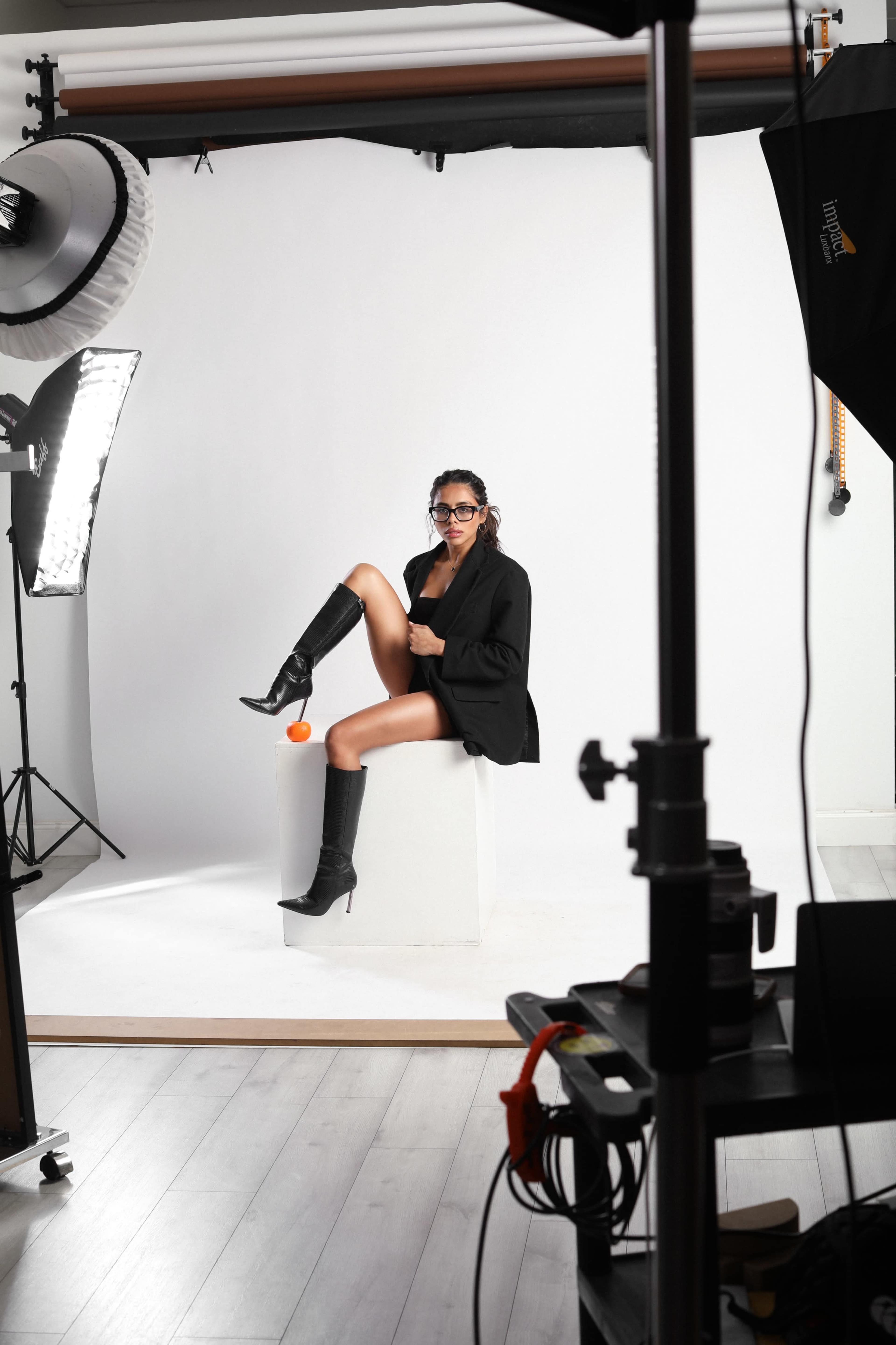 A model poses on a pedestal wearing a black blazer, knee-high boots, and glasses in a photography studio with lighting equipment.