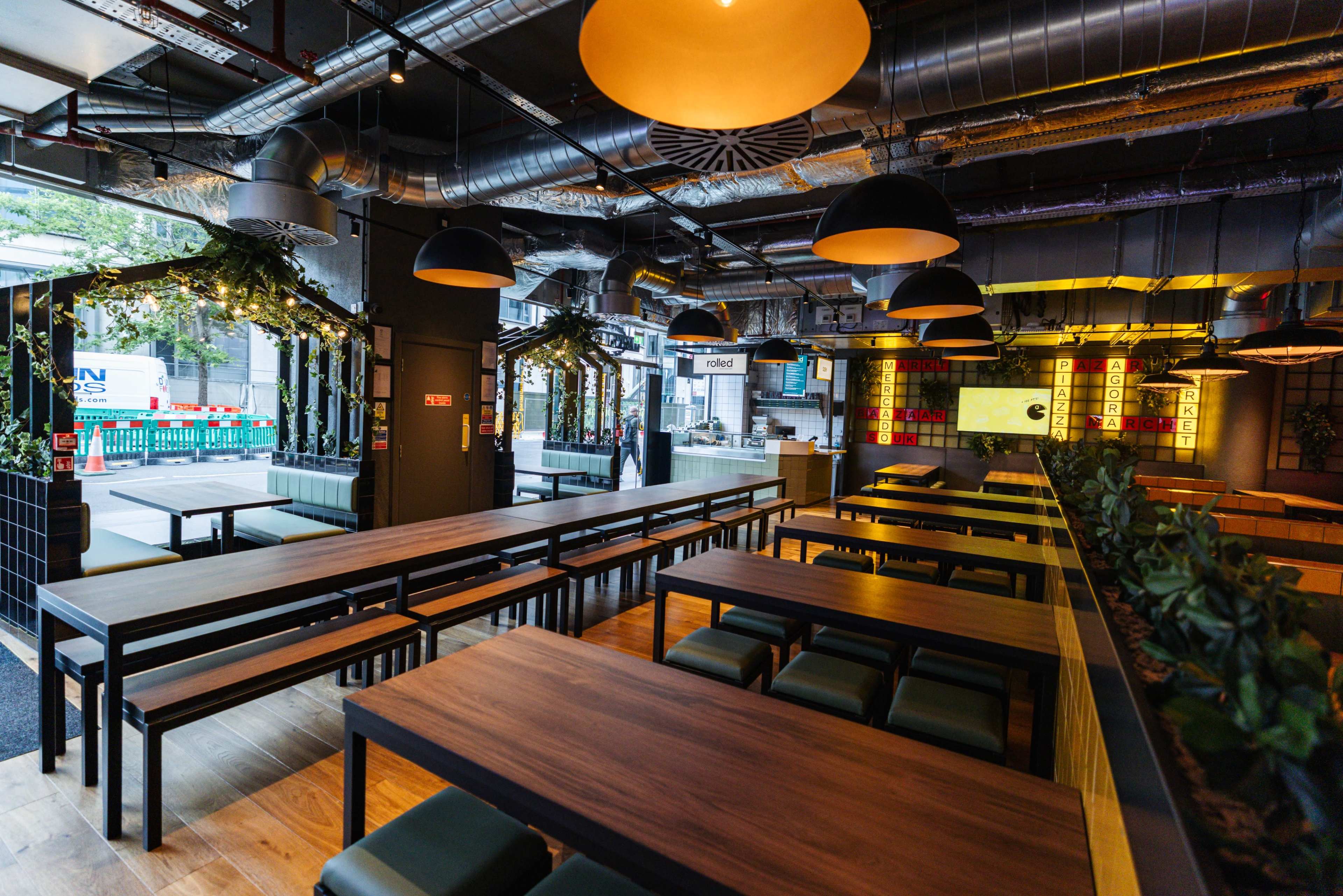 The image shows a modern restaurant interior with wooden tables, green seating, and industrial-style lighting fixtures.