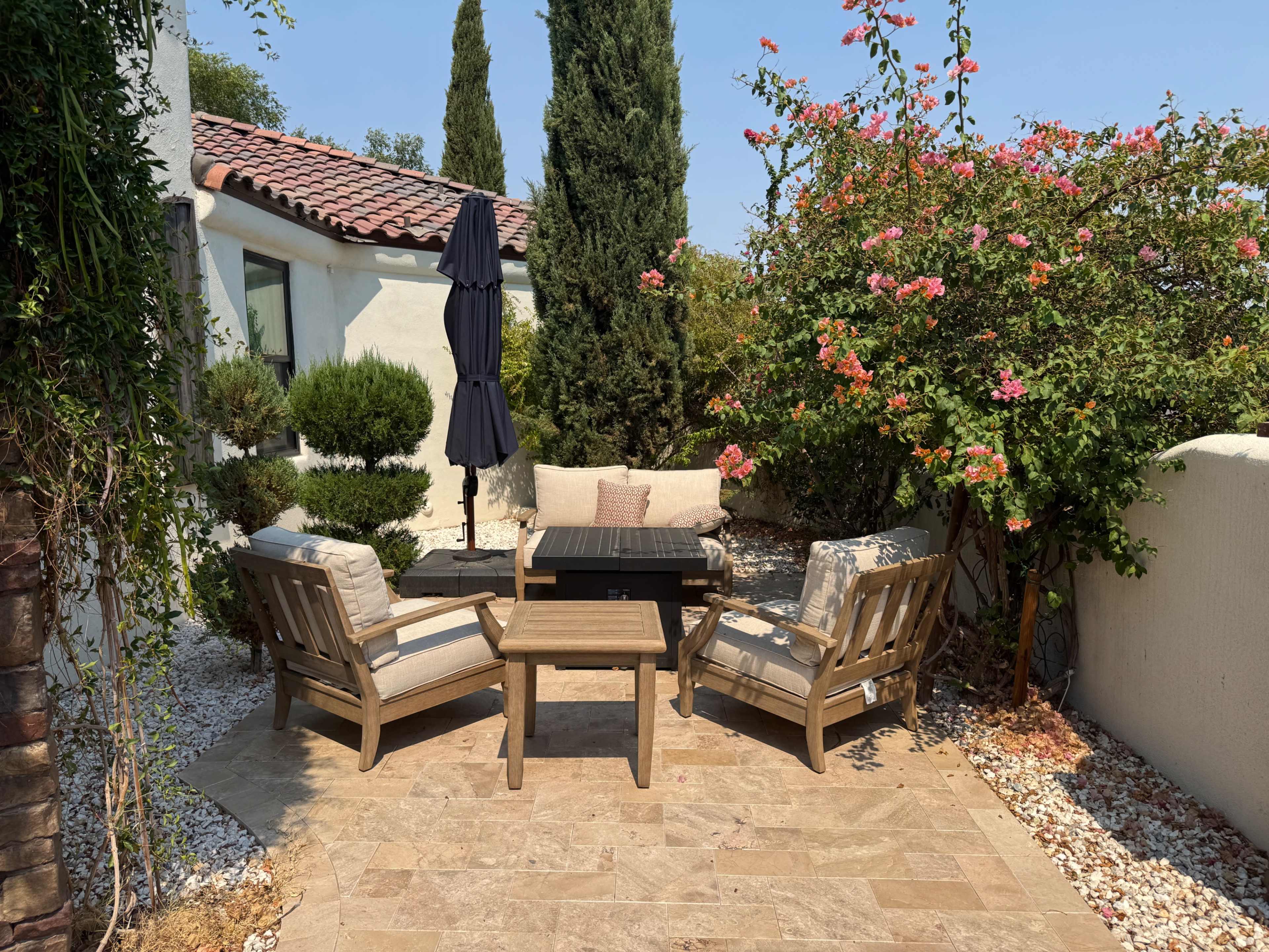 A small outdoor seating area features two wooden chairs and a table, surrounded by flowering plants and a tall umbrella.