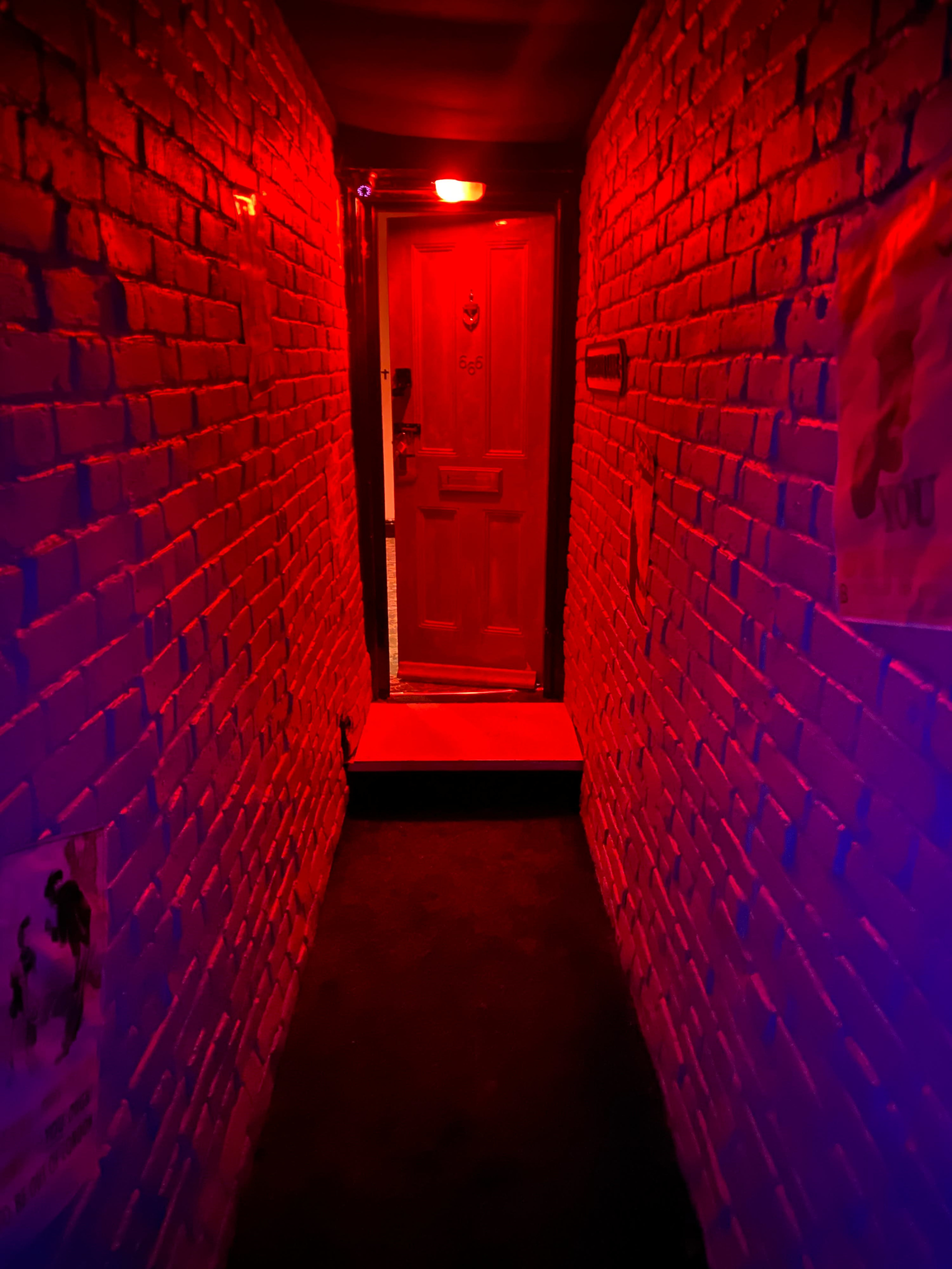 A narrow corridor with red-lit brick walls leads to a wooden door at the far end.