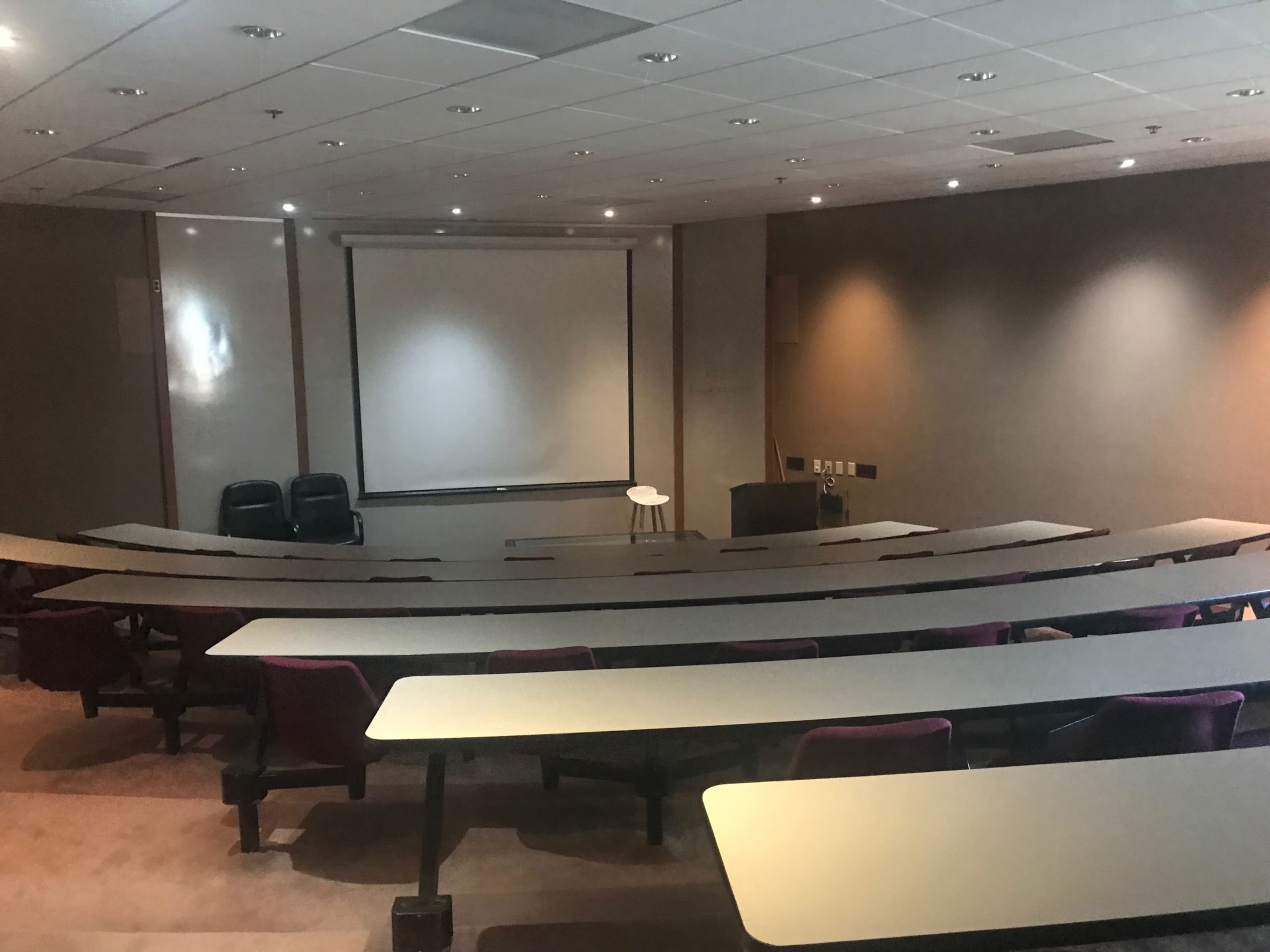 A lecture hall with tiered seating, a blank projection screen, and a small podium at the front.