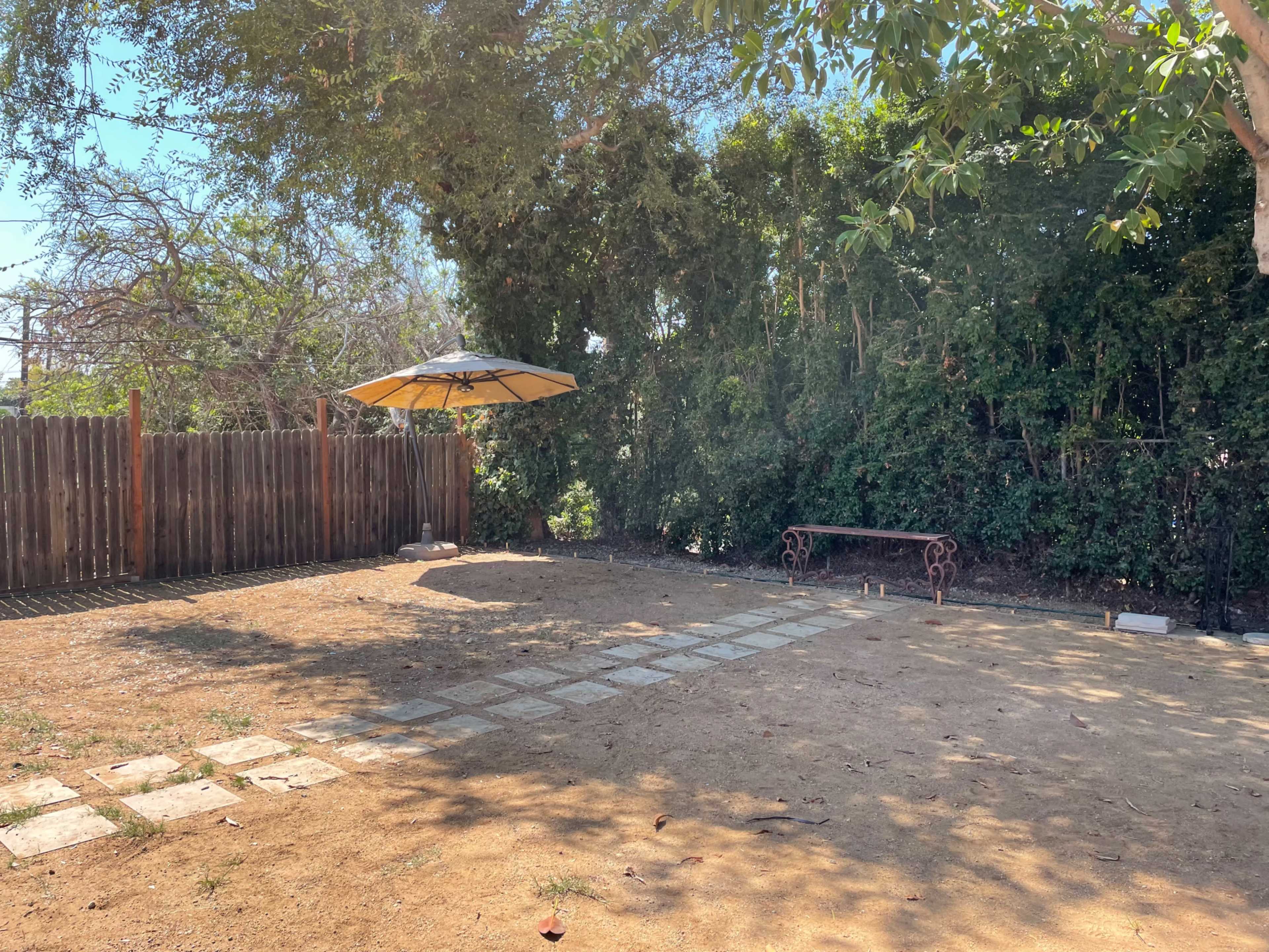 A bare, dirt backyard features a pathway of stone slabs leading to a bench under a large umbrella, surrounded by greenery and a wooden fence.