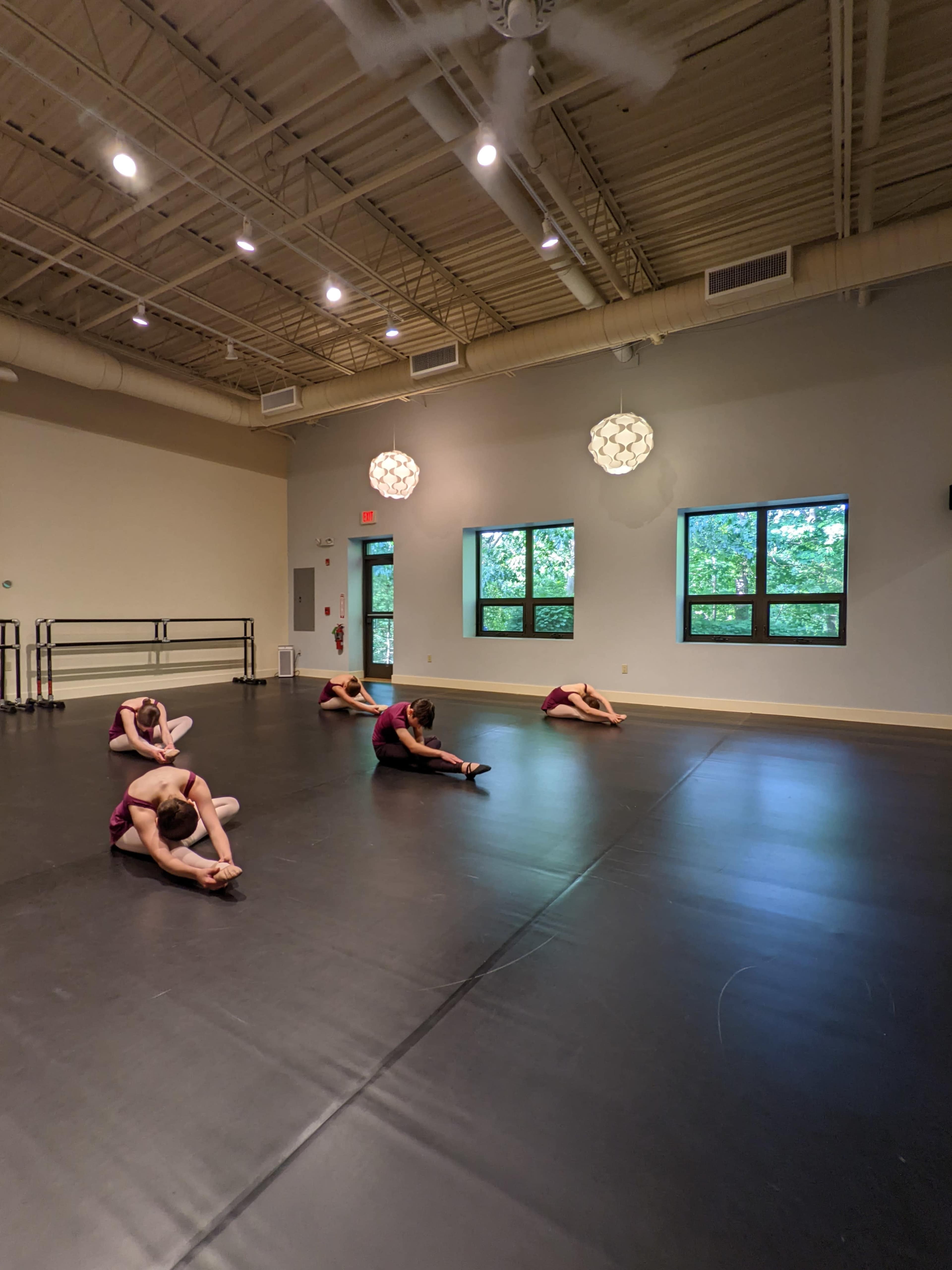A group of dancers is stretching on a black mat in a brightly lit studio with large windows.