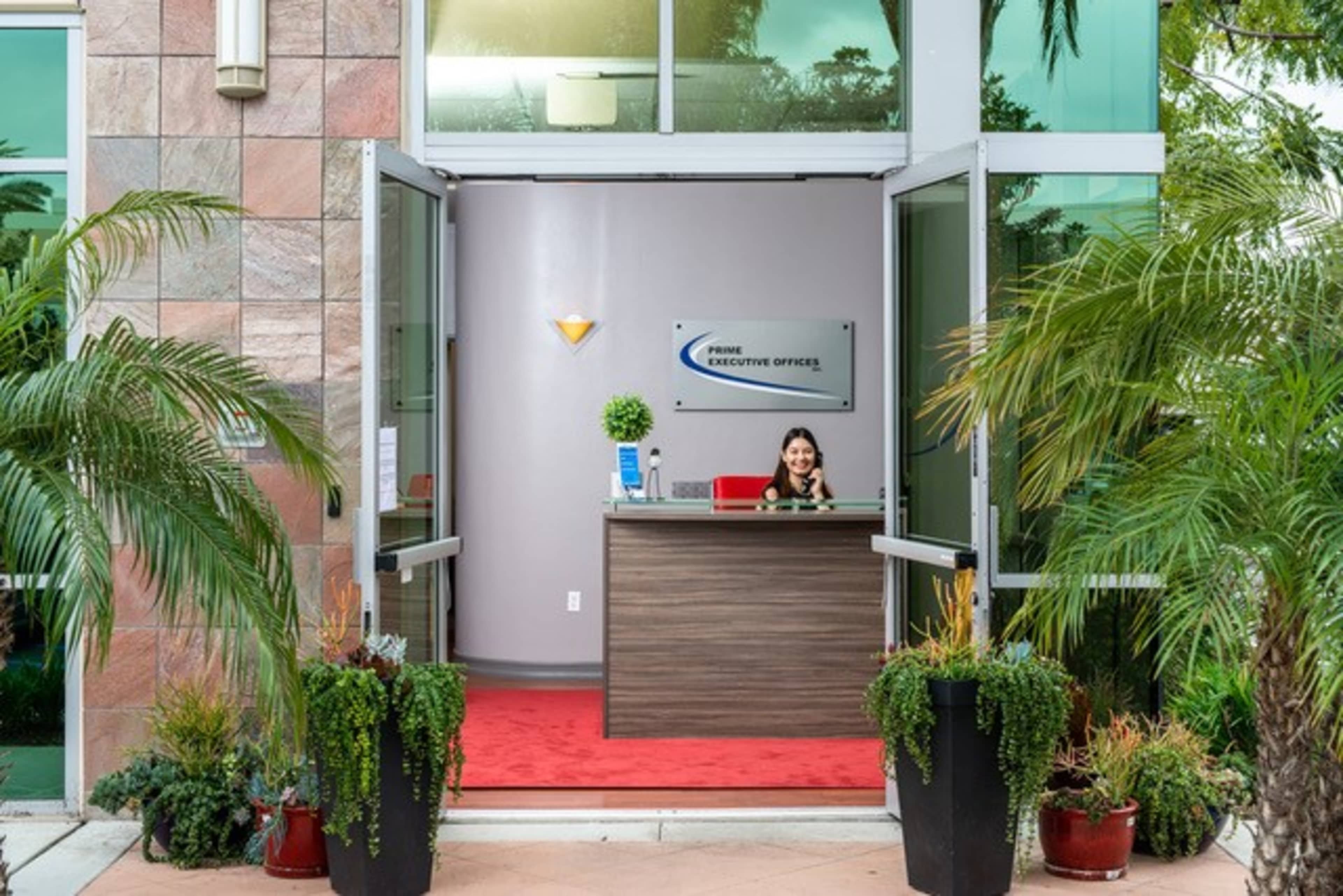 A receptionist smiles from behind a wooden front desk in a modern office lobby surrounded by potted plants.