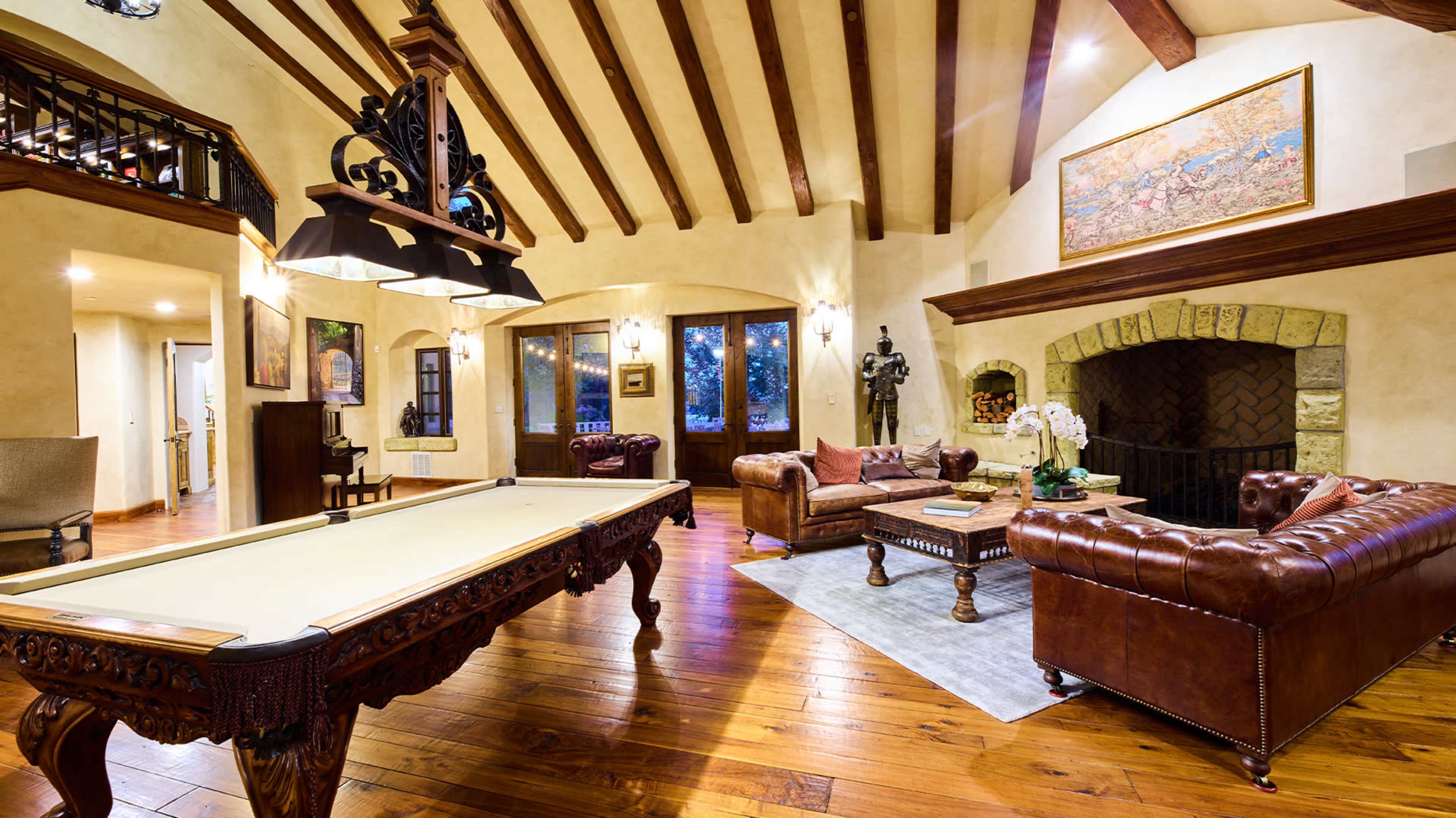 A spacious living area with a pool table, leather sofas, and a stone fireplace, featuring wooden beams and warm-toned furnishings.