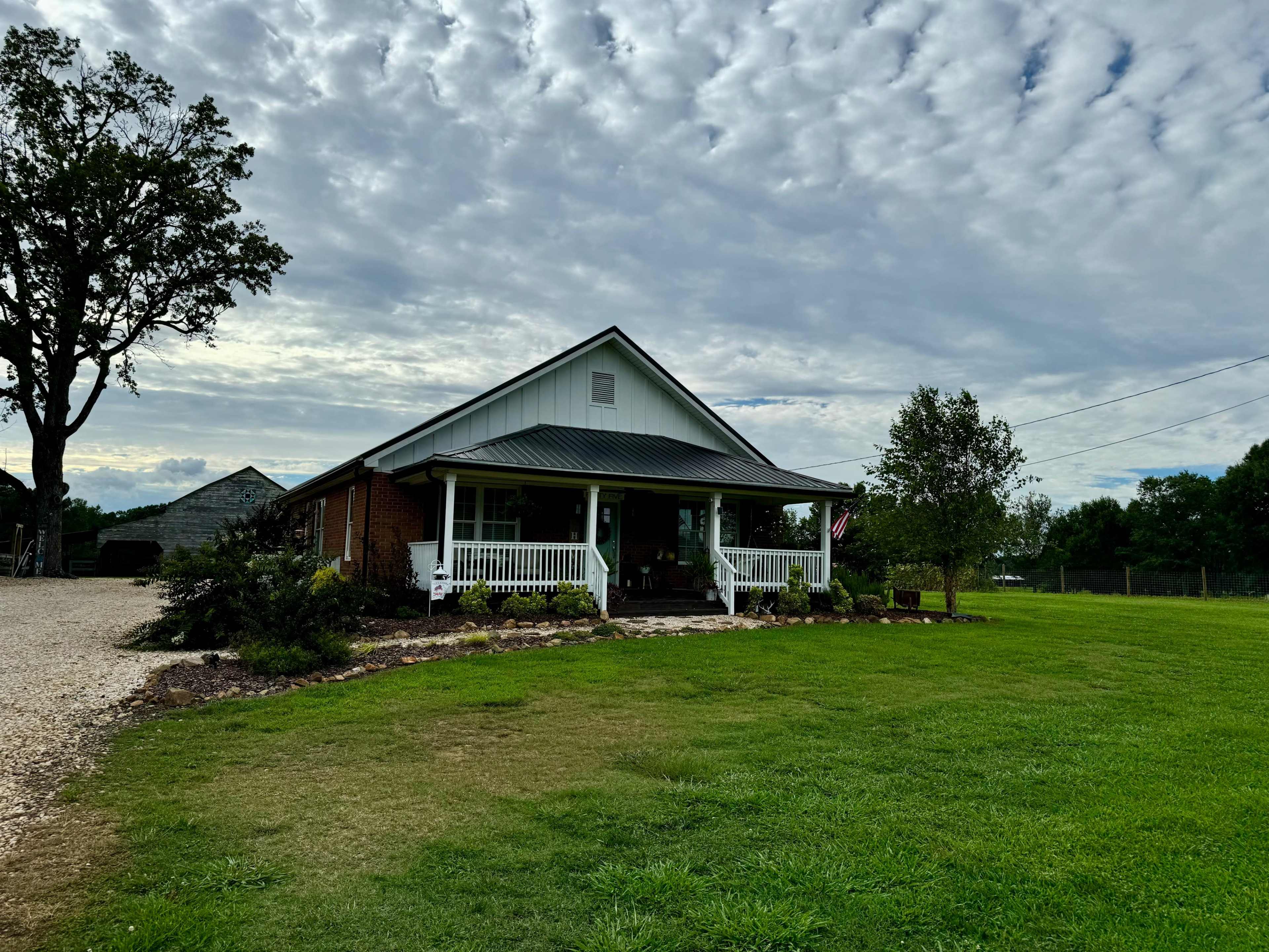 A single-story house with a metal roof and front porch is surrounded by a grassy lawn and trees under a cloudy sky.