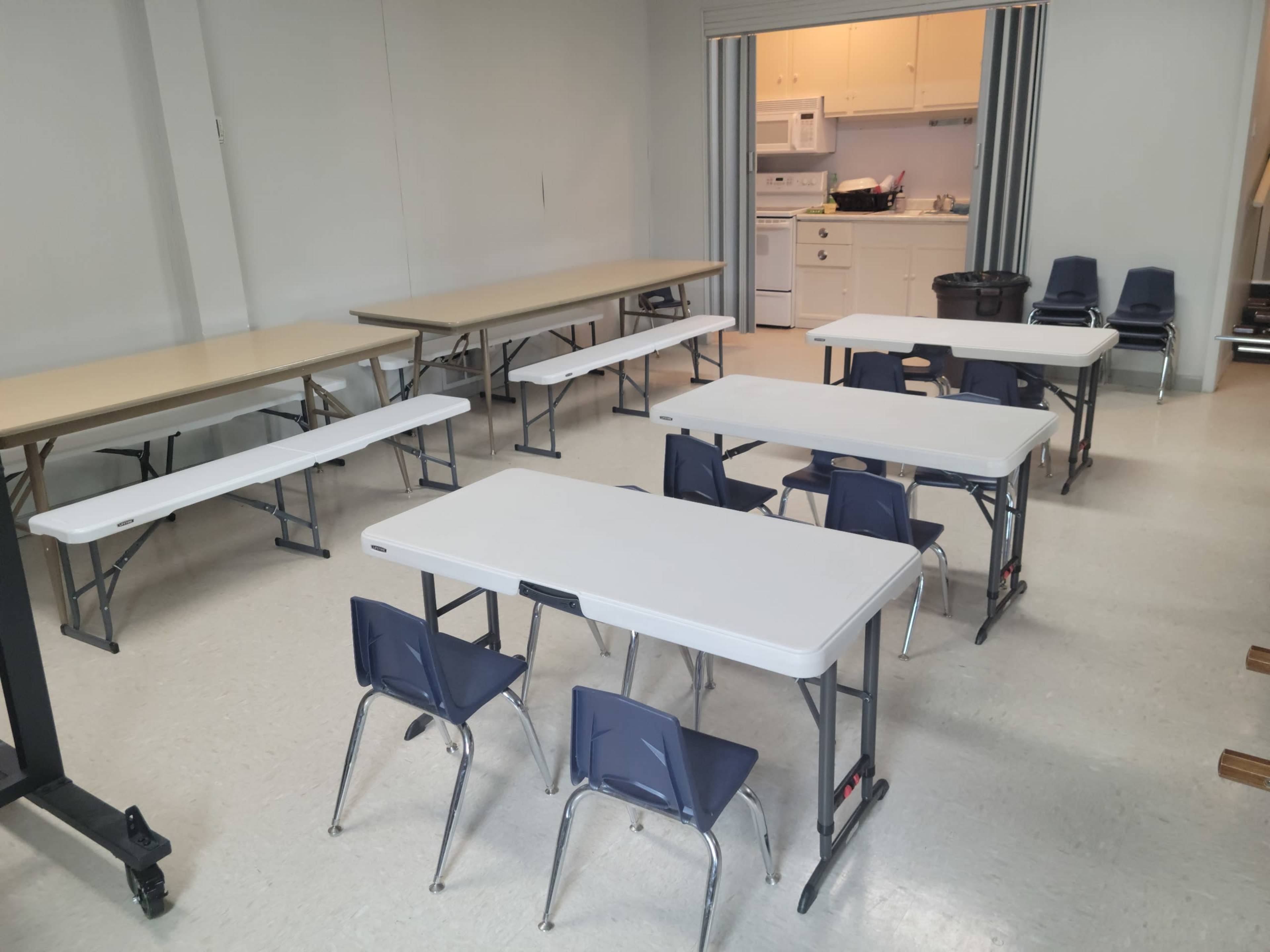 A classroom setup with several folding tables arranged in rows and blue chairs positioned around them, with a kitchenette area visible in the background.