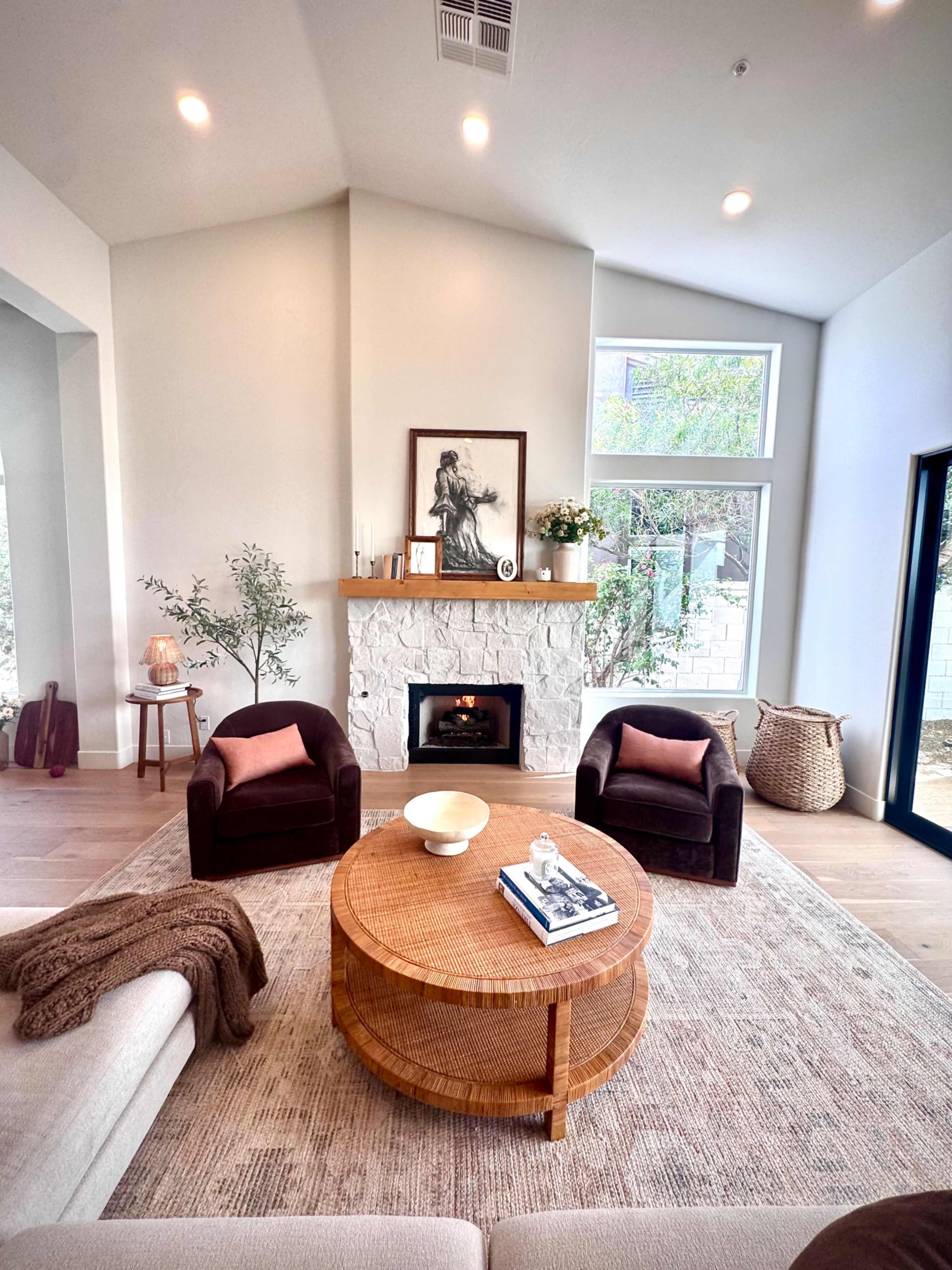 The image shows a modern living room featuring a round coffee table, two armchairs, a stone fireplace, and large windows allowing natural light to enter the space.