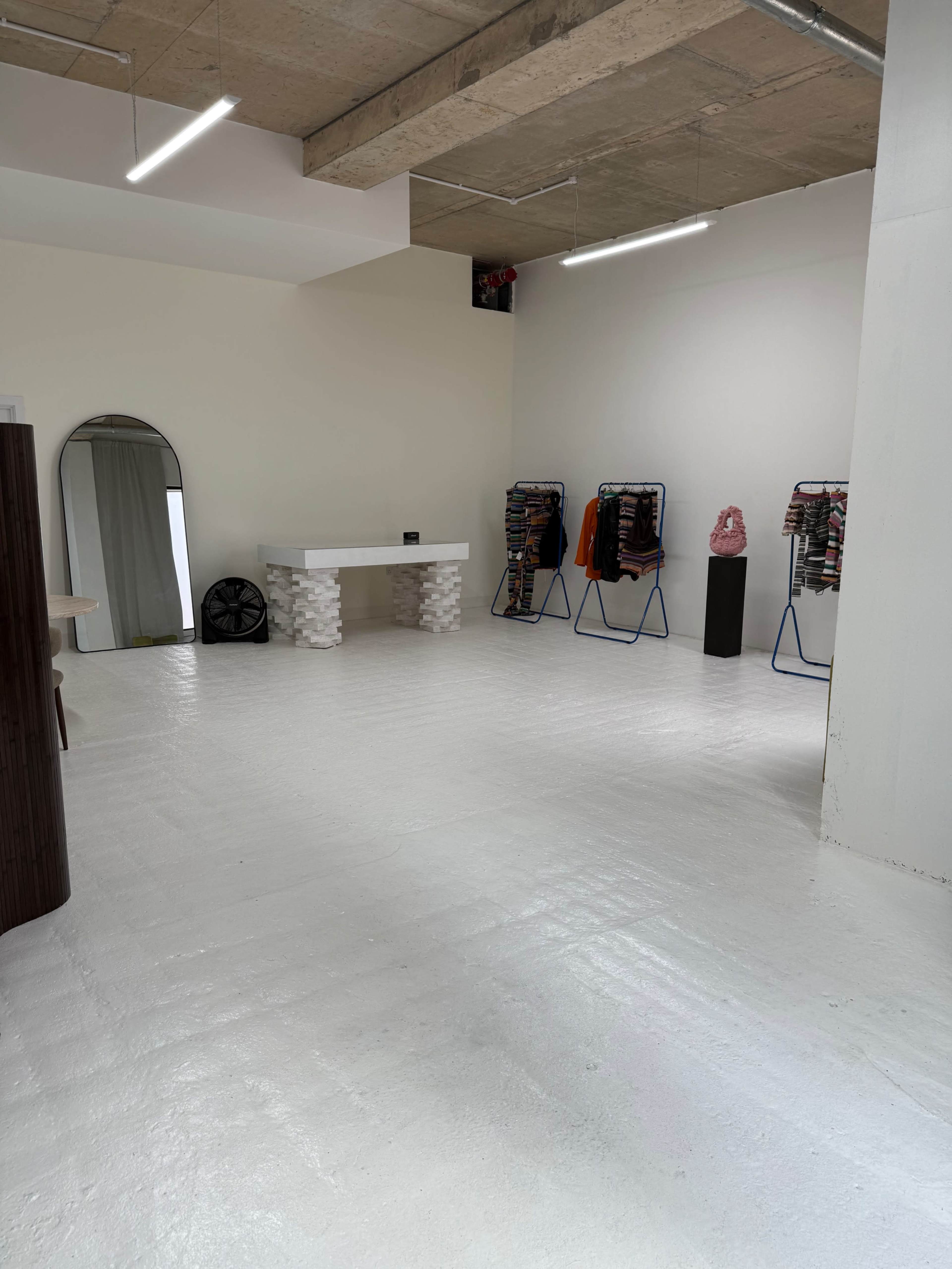 The image shows a spacious, brightly lit interior with a white floor, a large mirror against the wall, clothing racks displaying garments, and various display stands.