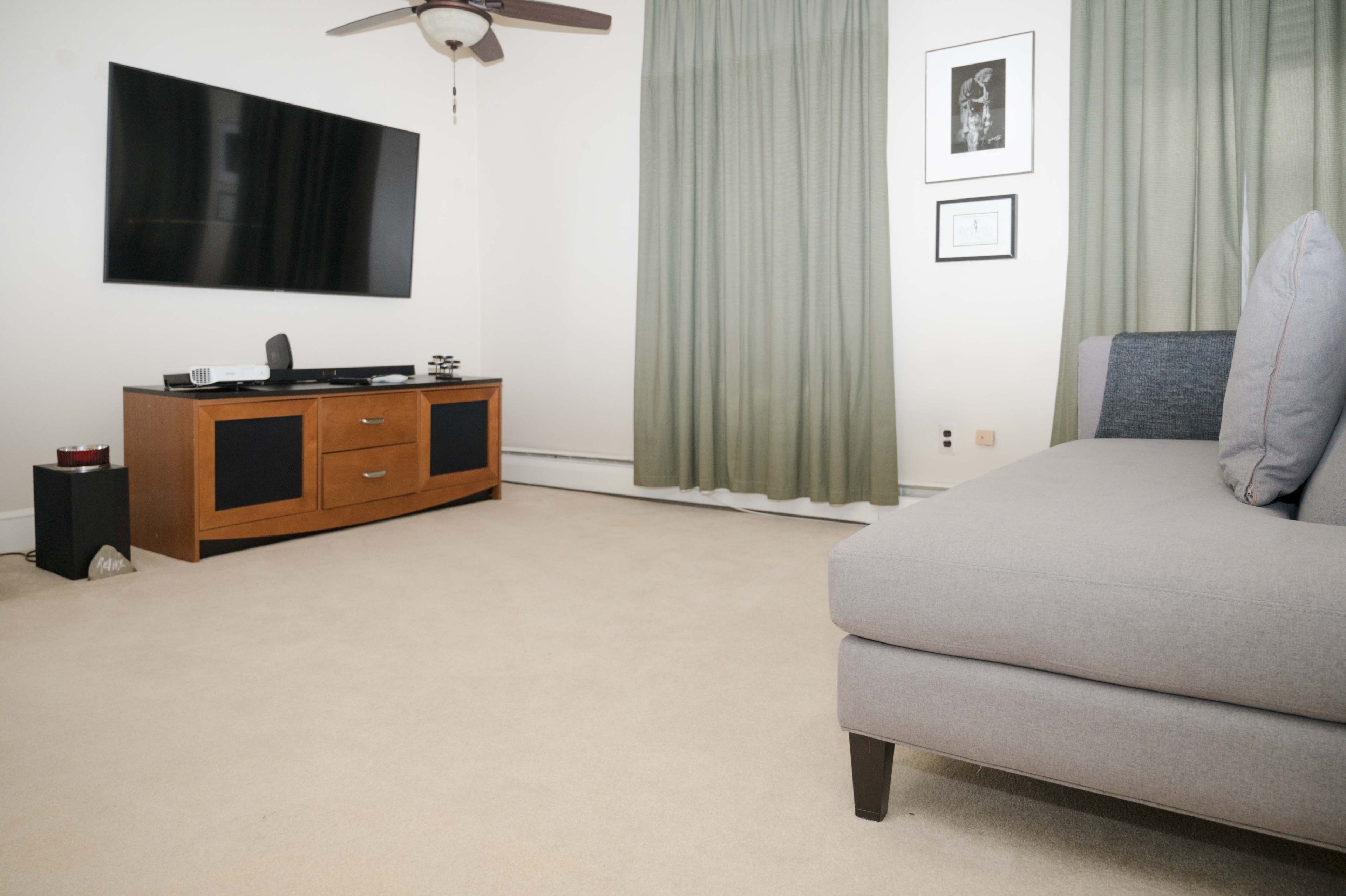 The image shows a living room with a television mounted on the wall, a wooden entertainment unit, and a gray sofa with cushions on a beige carpet.
